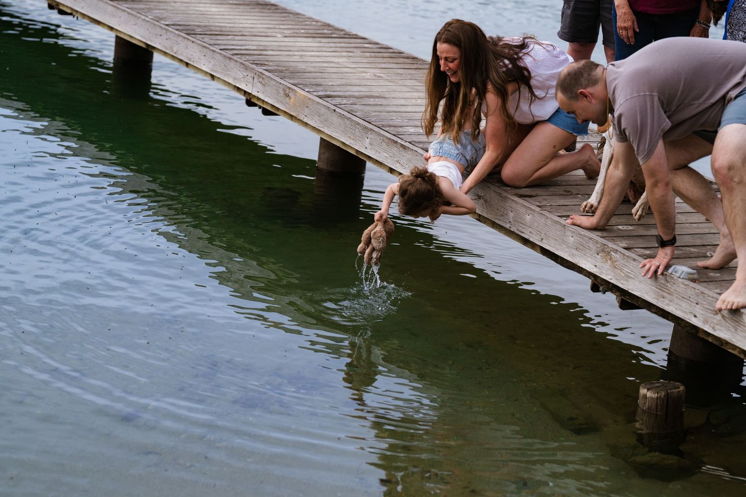 People sitting on a wooden dock reaching into green lake water on a sunny day.