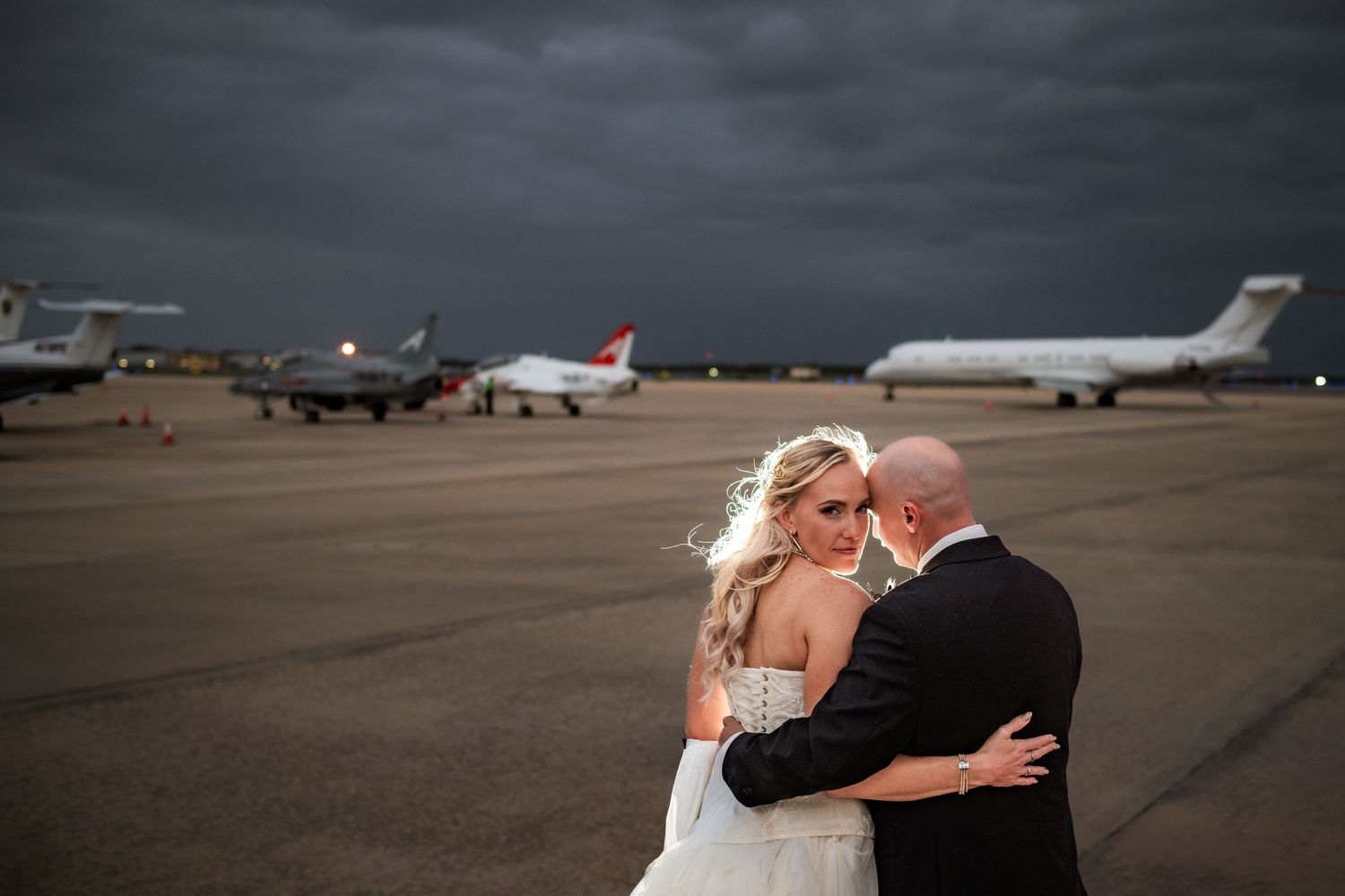 Bride and groom embrace romantically on airport tarmac with jets and dramatic stormy sky backdrop.
