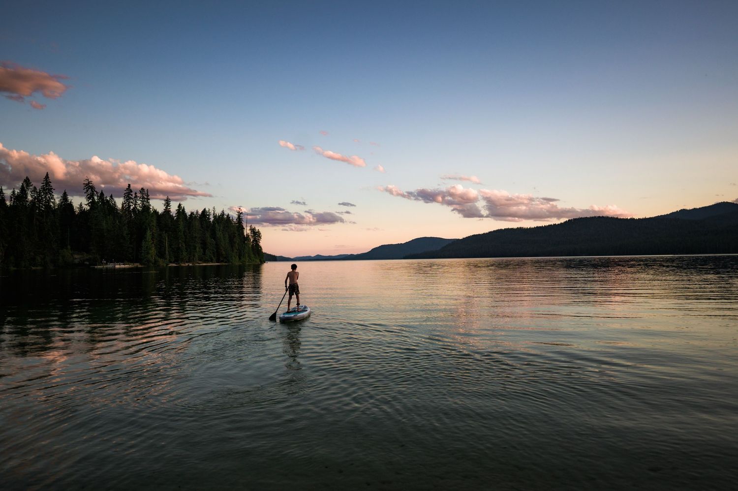 Panoramic view of a paddle boarder silhouetted against a tranquil lake at sunset with mountains in the background.