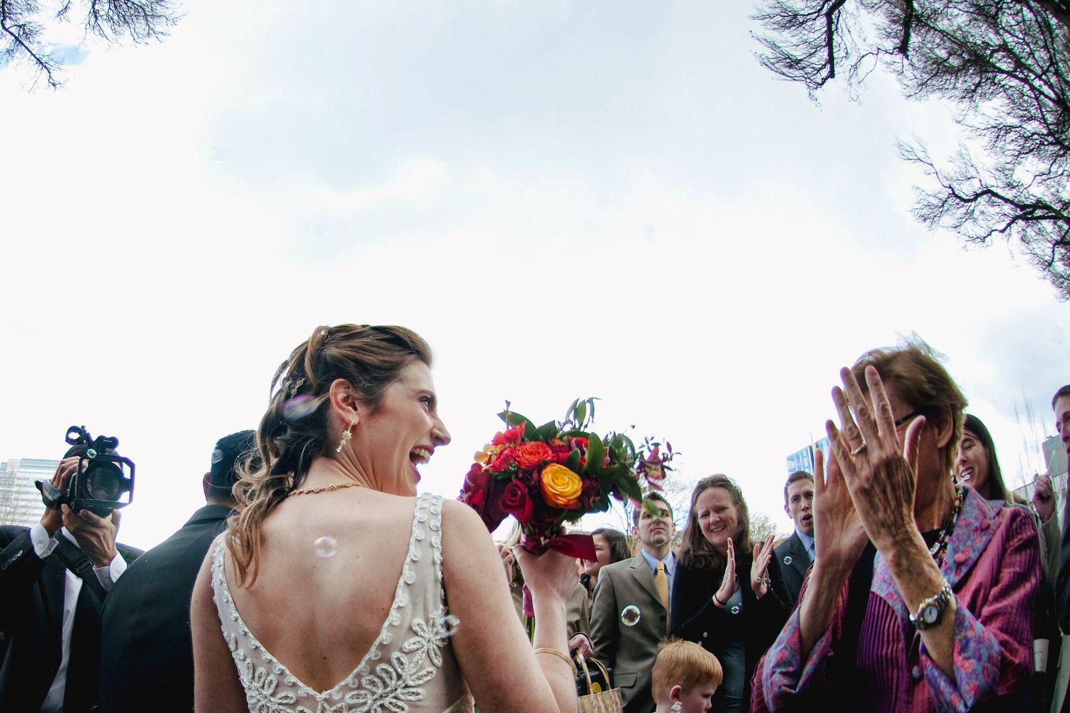 Bride in lace gown holds colorful bouquet as guests clap and celebrate at an outdoor wedding ceremony.