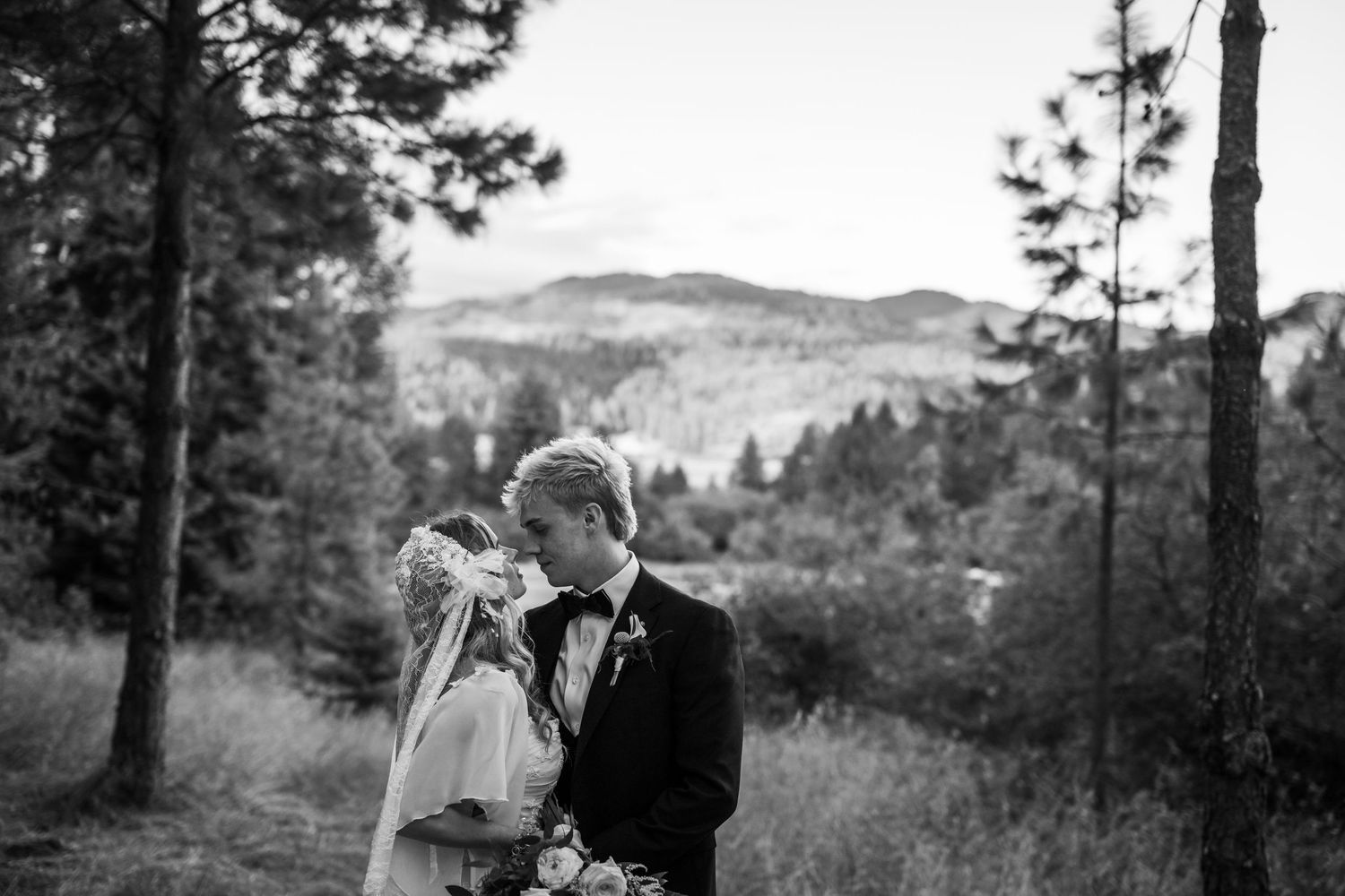Black and white portrait of a bride and groom sharing a romantic moment surrounded by pine trees and mountains.