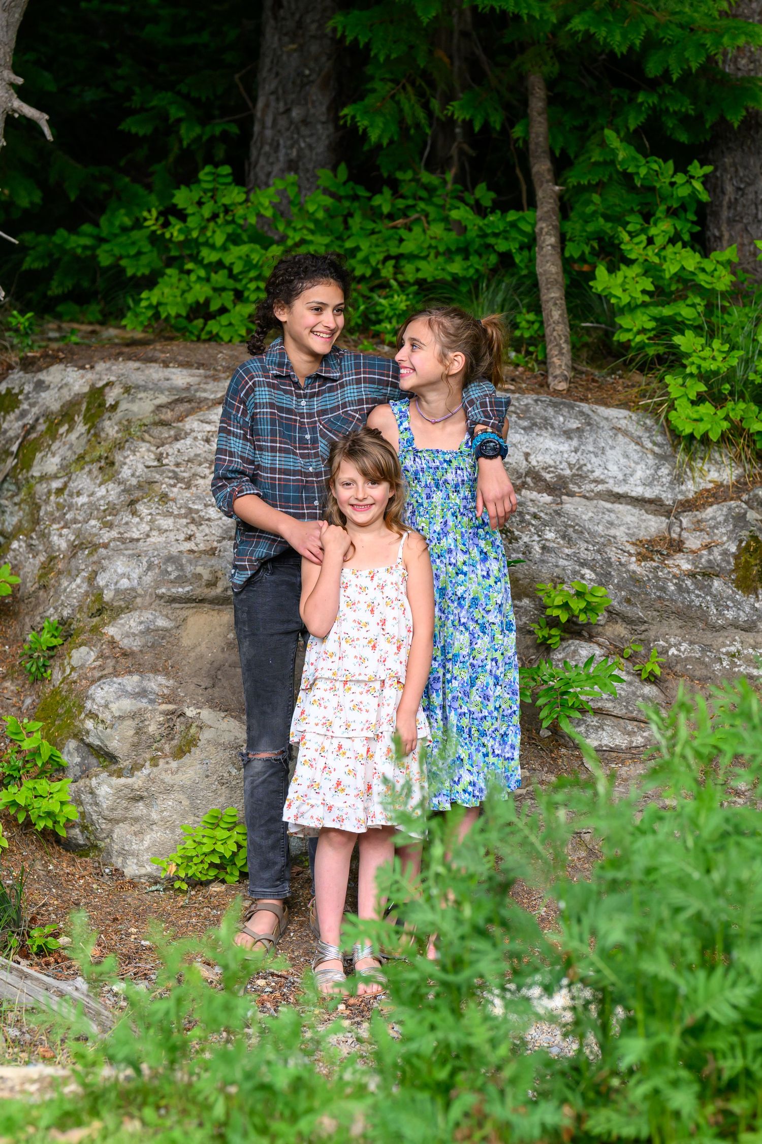 Three smiling girls posing together on rocky terrain surrounded by lush green forest trees.
