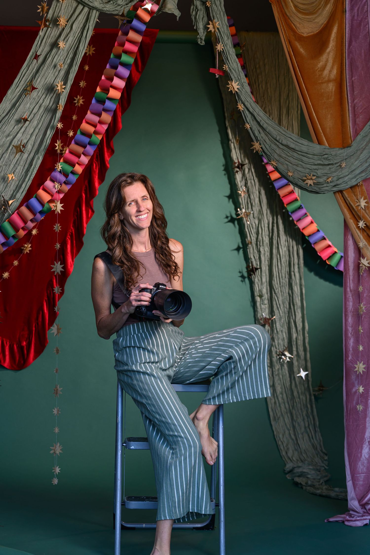 Smiling woman photographer sitting on stool holding camera in colorful studio with draped fabric and paper chain decorations.