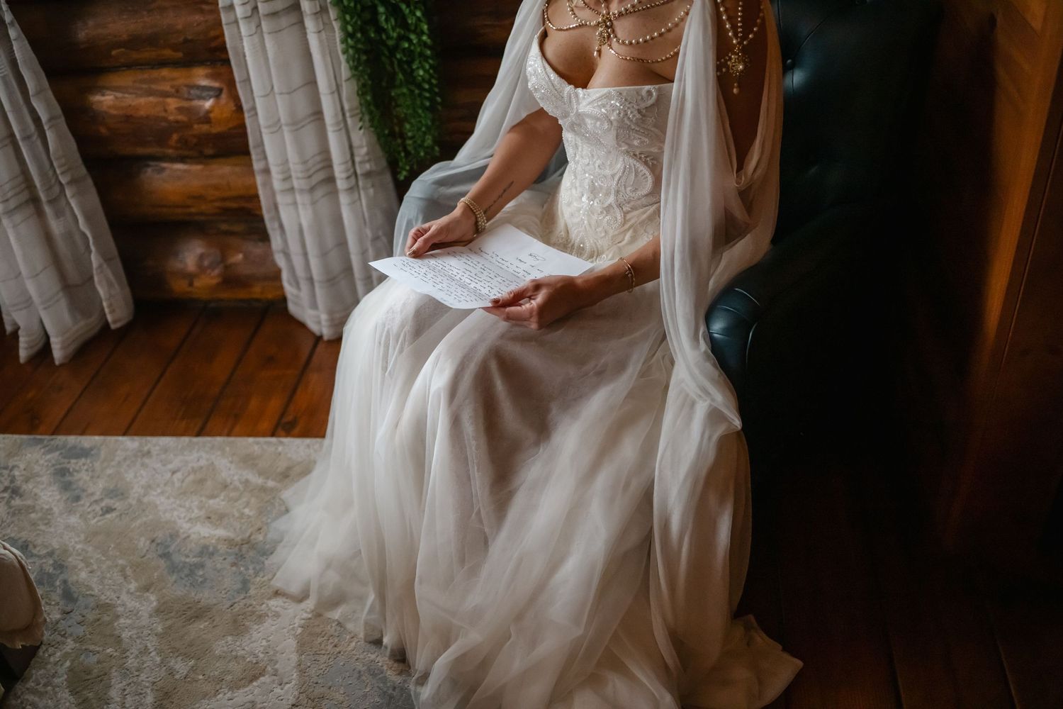 Bride in lace wedding gown reading vows letter while seated in rustic log cabin interior.
