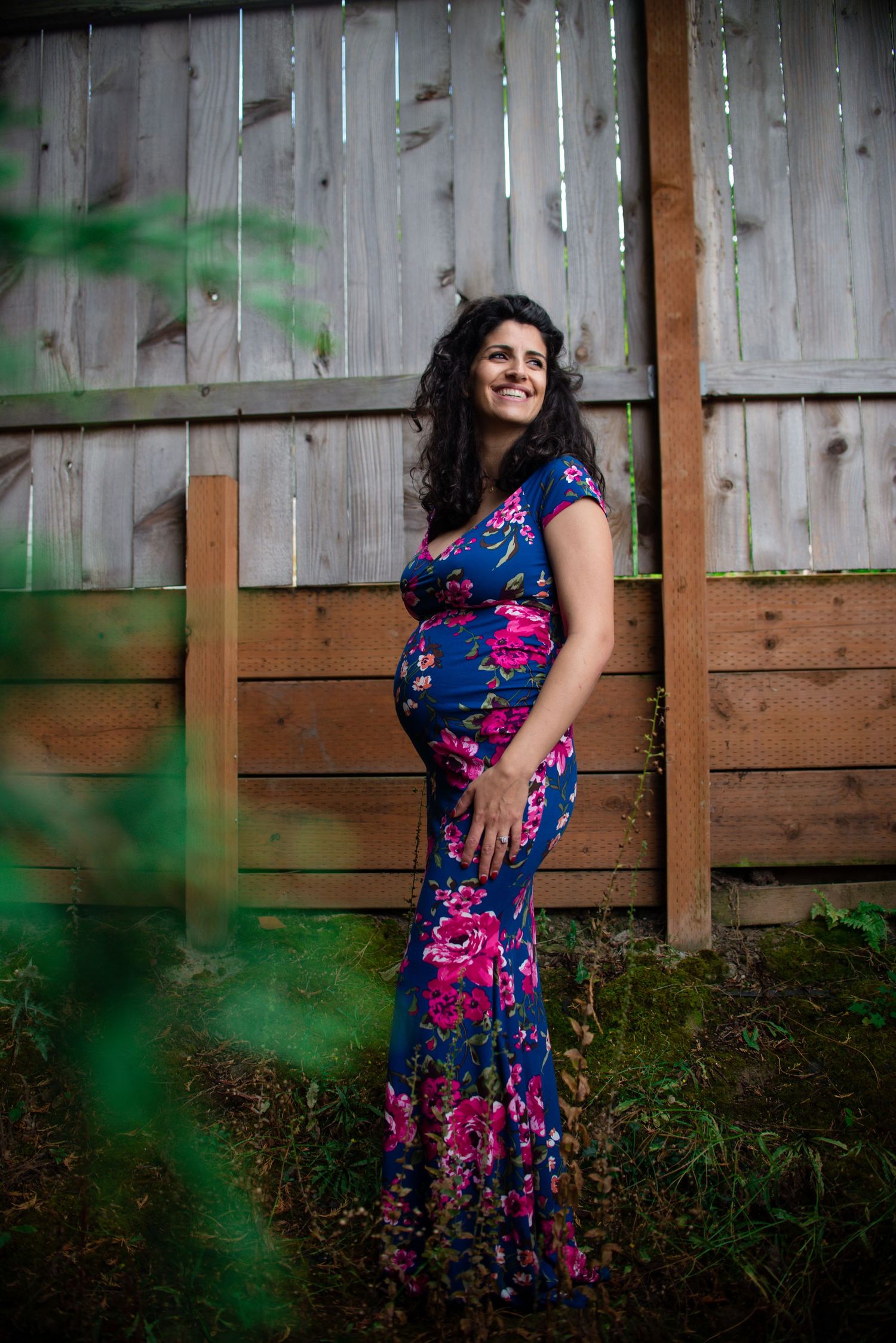 A woman in a vibrant blue and pink floral dress poses against a wooden fence in a garden setting.