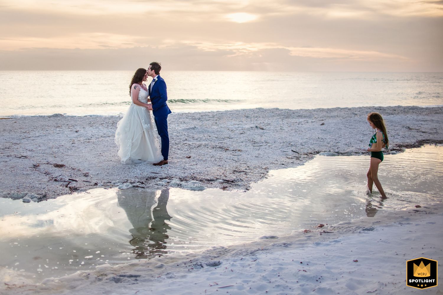 A coastal sunset scene with a couple in wedding attire by the water's edge while a child plays nearby on the beach.