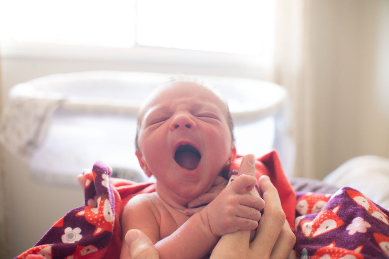 A newborn baby in red and white clothing yawns while resting on a white surface.