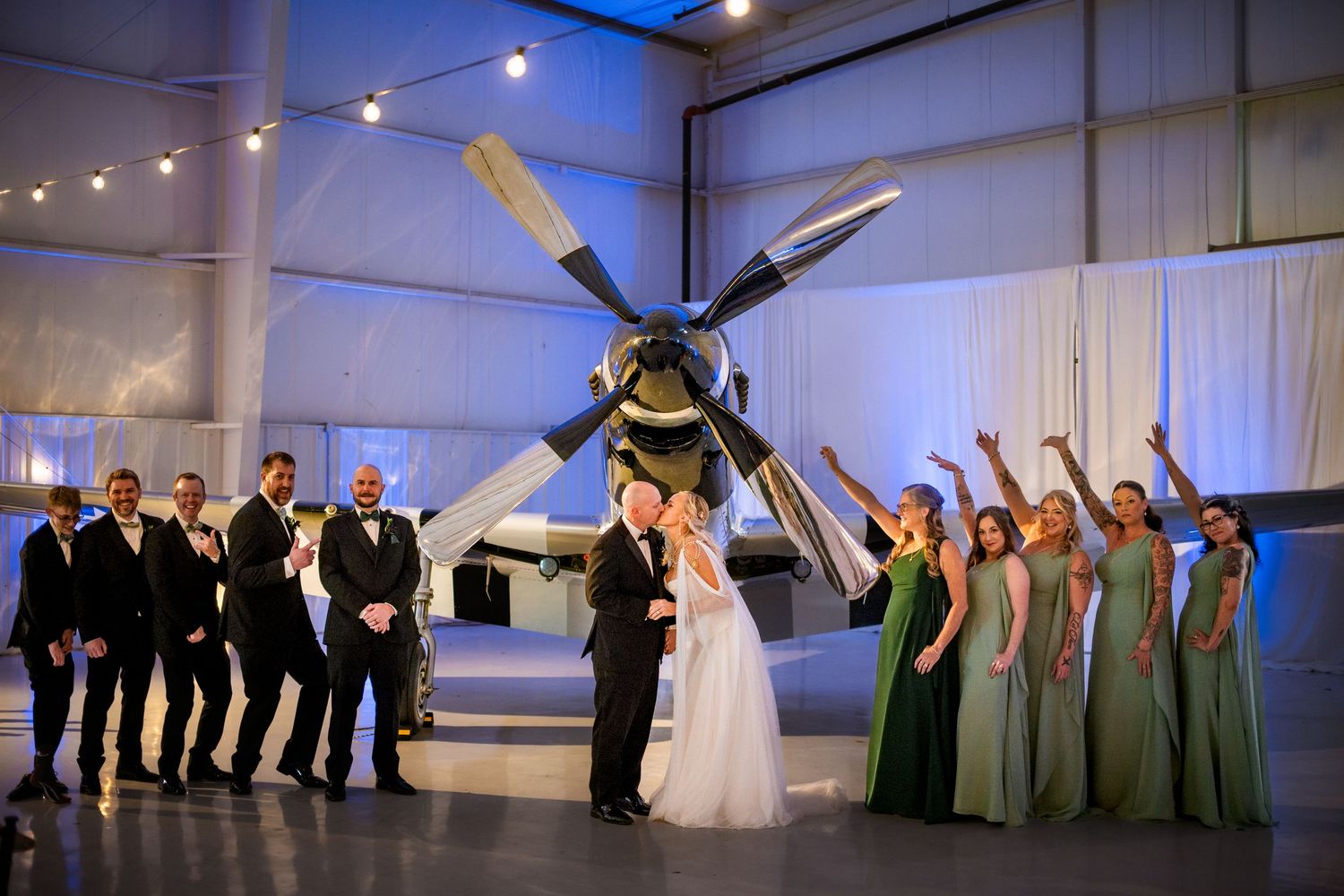 Wedding party poses before vintage airplane in hangar venue with bridesmaids in green gowns.