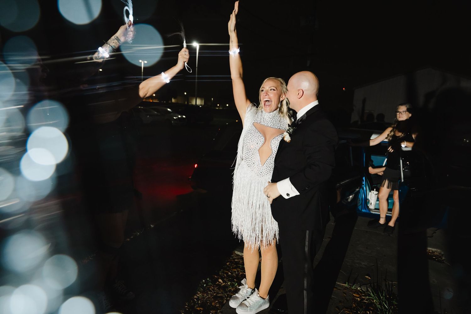 Bride in white fringe dress celebrates with groom at night wedding reception, raising arm joyfully.