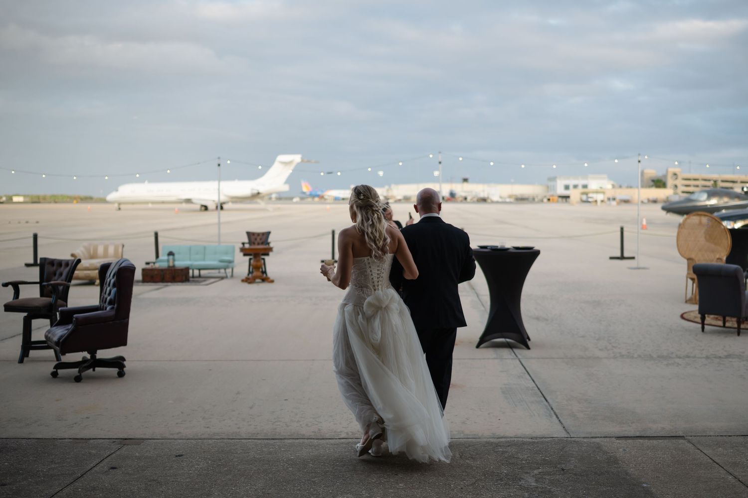 Bride and groom overlook airport tarmac with private jet during outdoor wedding cocktail hour.
