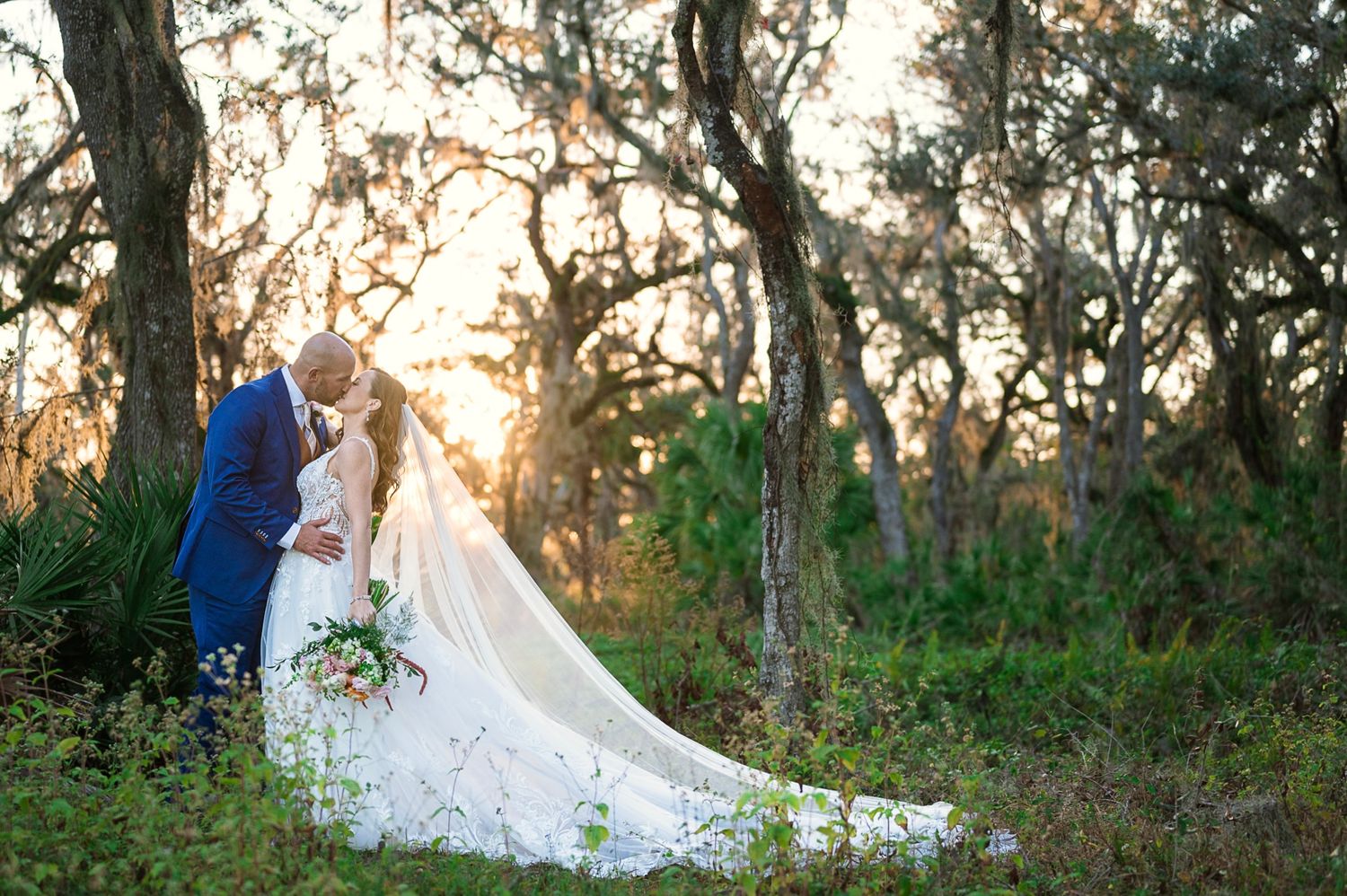 Couple shares romantic moment in white wedding attire at sunset among trees in a natural outdoor setting.