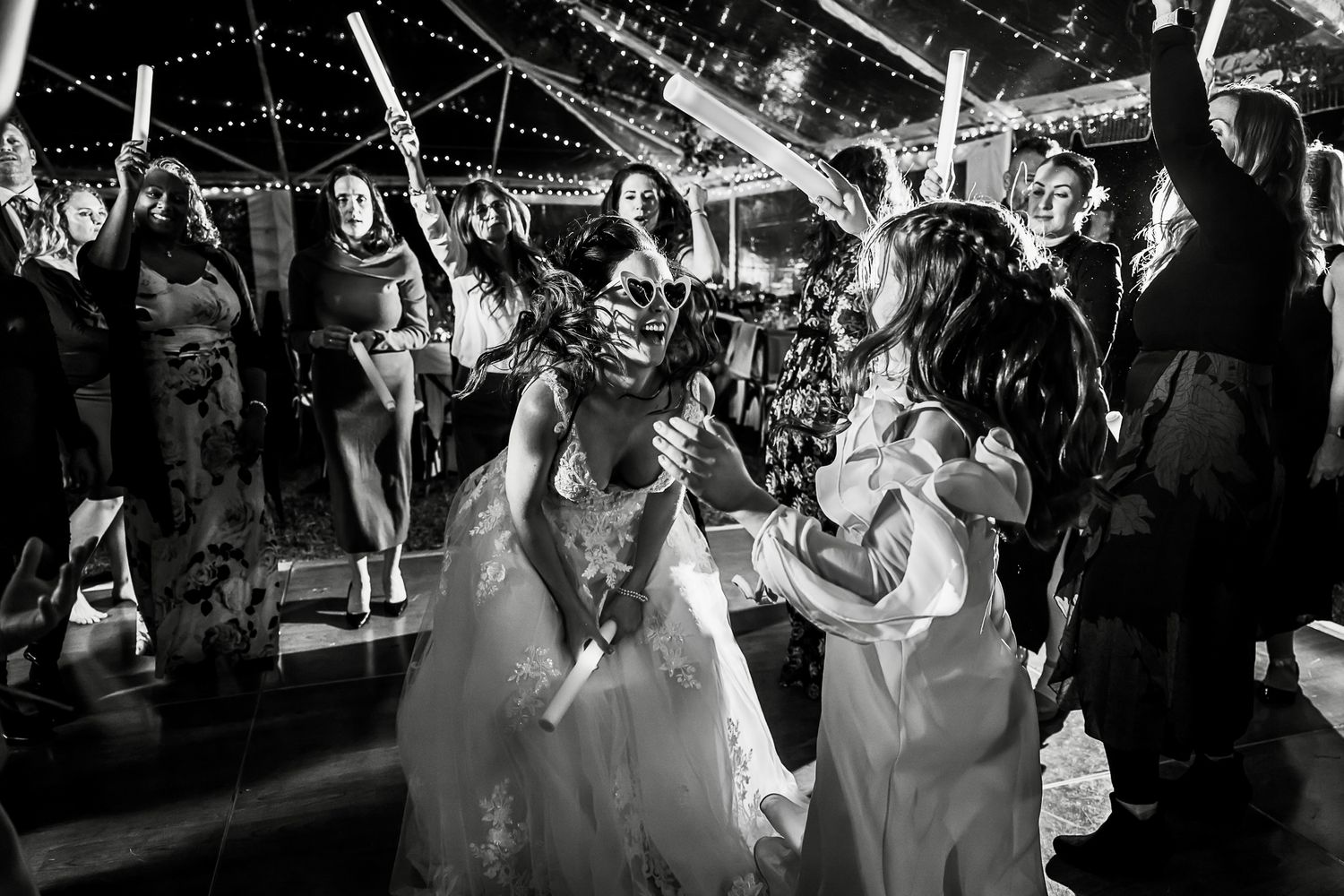 Black and white photo of two brides dancing together at their wedding reception under twinkling lights and celebrating guests.