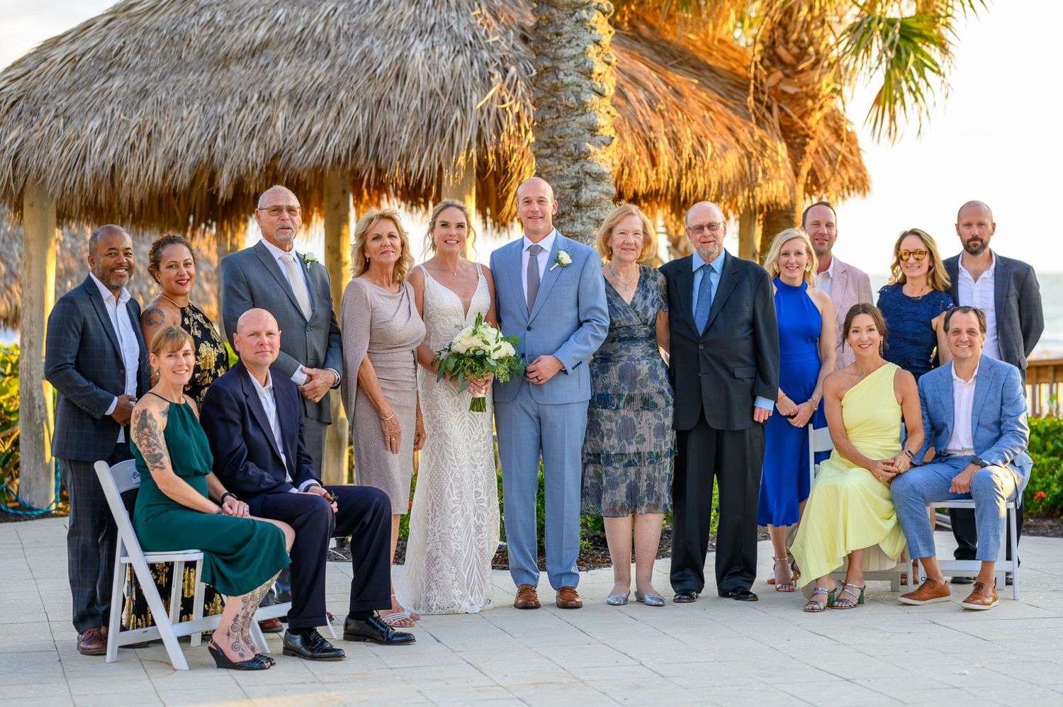 Group photo at a beach wedding under a thatched palm roof pavilion.