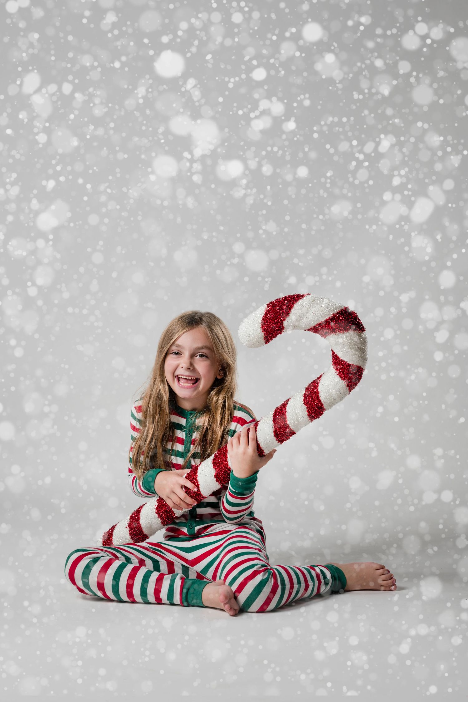 Child wearing red and white striped holiday pajamas poses playfully on gray background with falling snow effect.
