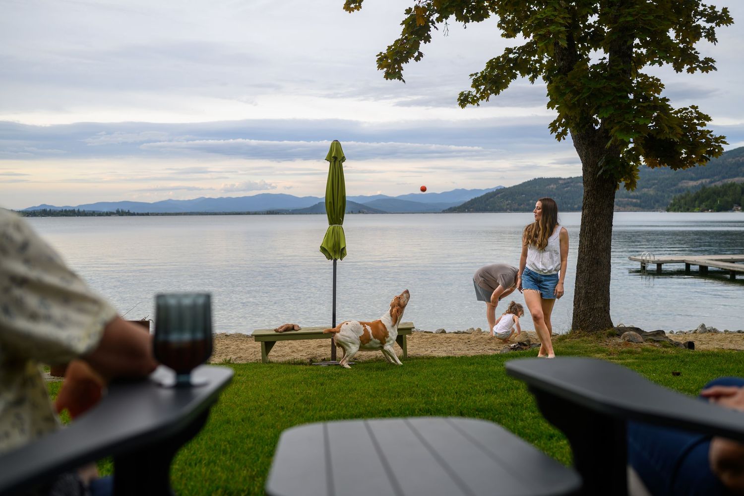 Family enjoying lakeside beach with dogs, green umbrella, and mountain views at dusk.