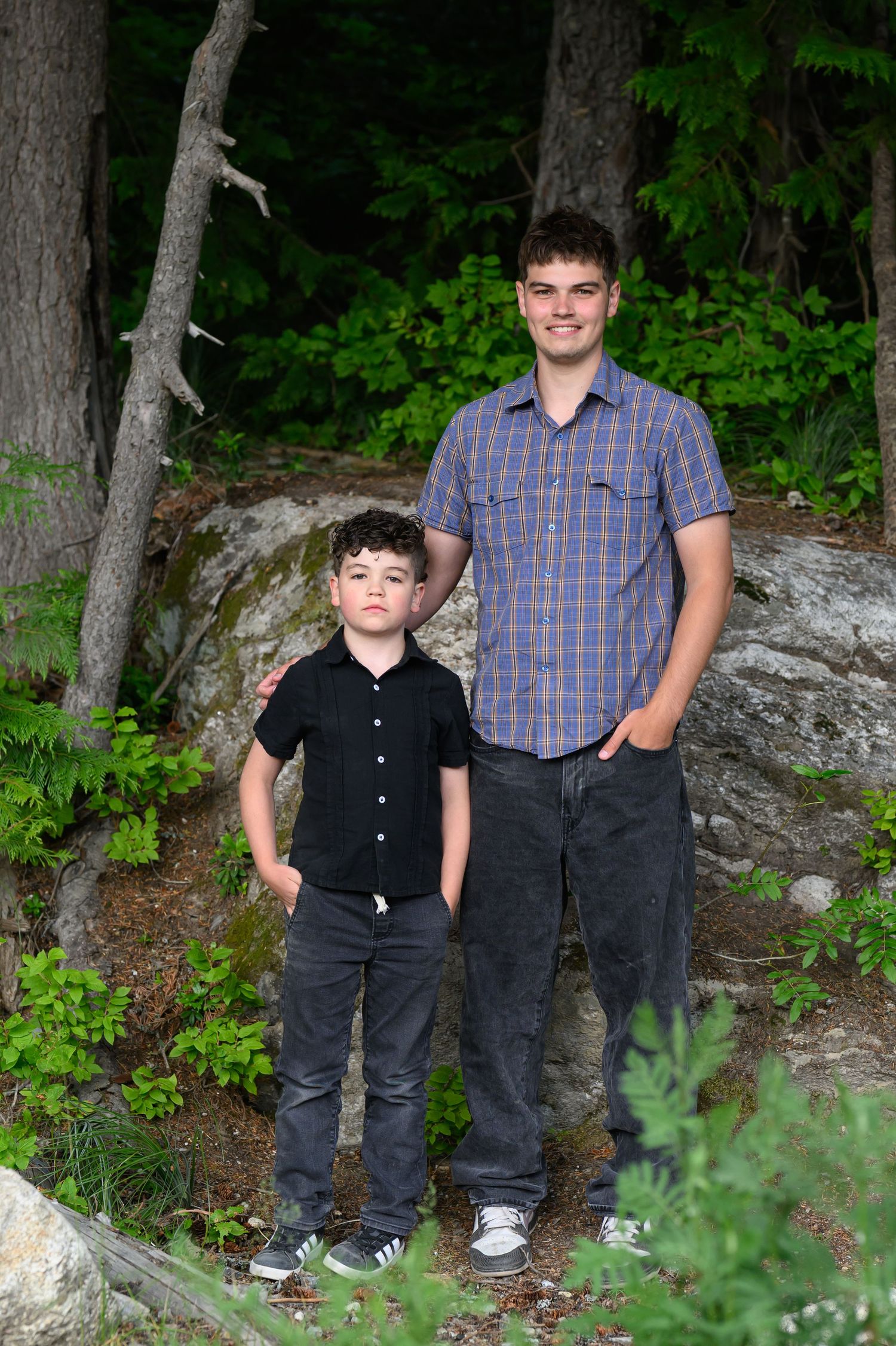 Two boys, one tall teen and one young child, pose together outdoors among rocks and green trees.
