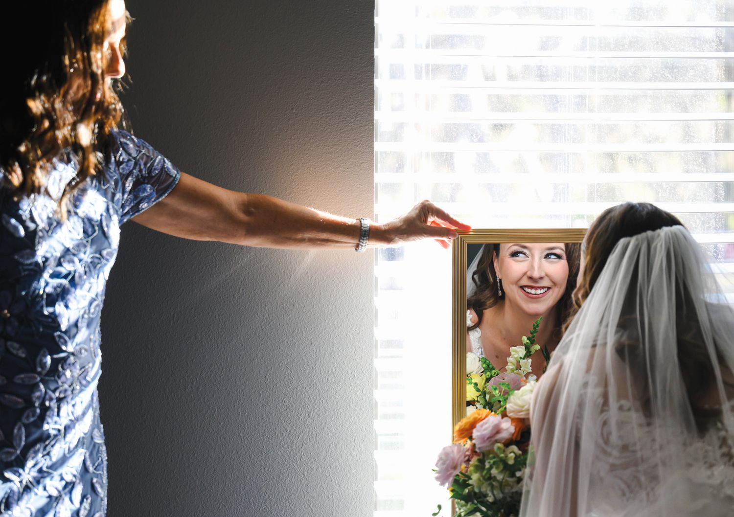 Bride in veil holding bouquet admires her reflection in mirror while woman in blue dress adjusts window blind.