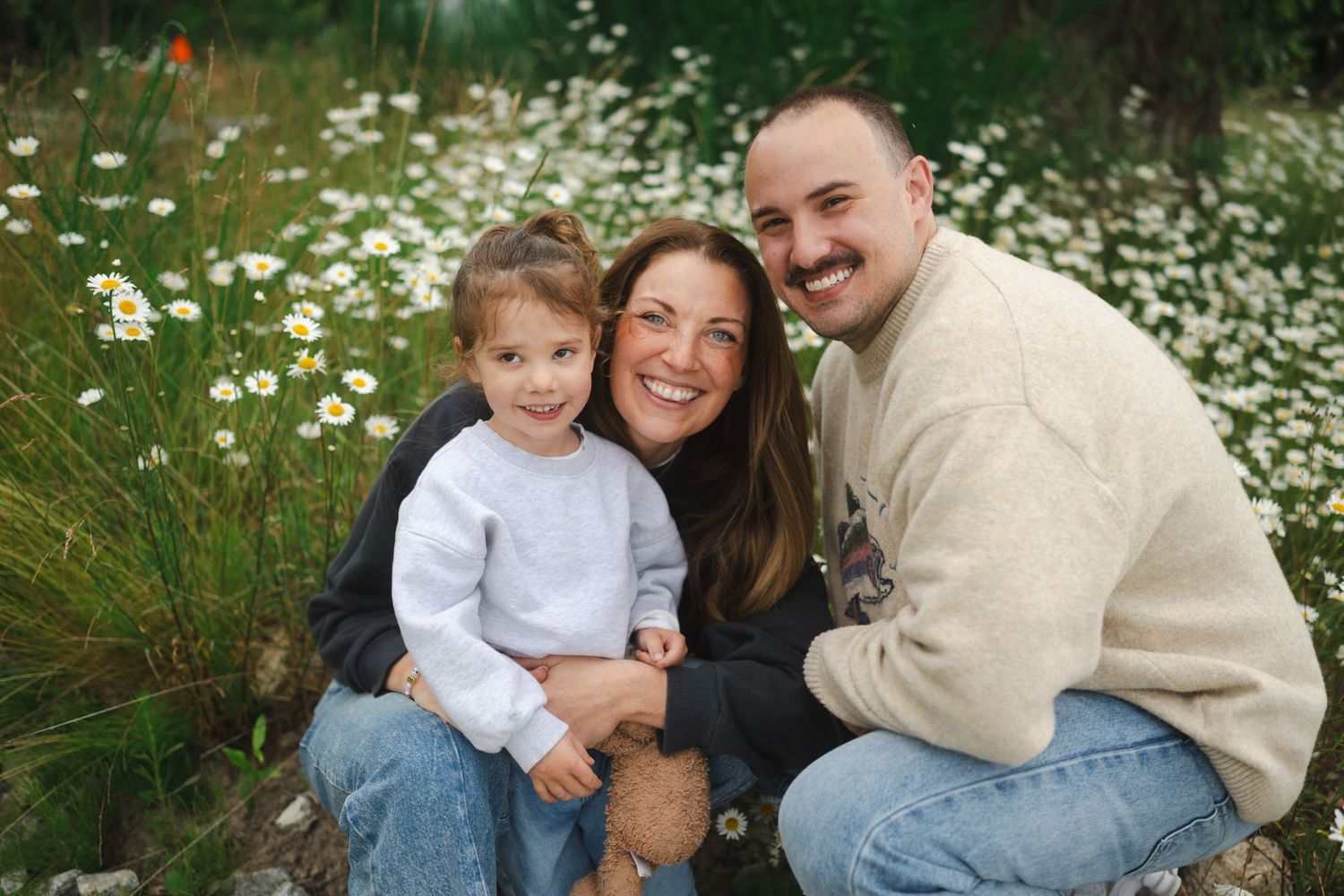 Family of three smiling together in a wildflower meadow with daisies blooming in the background.
