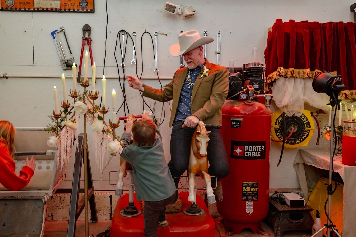 Man in cowboy hat shows child a toy horse in a workshop with tools and a red Porter Cable compressor.