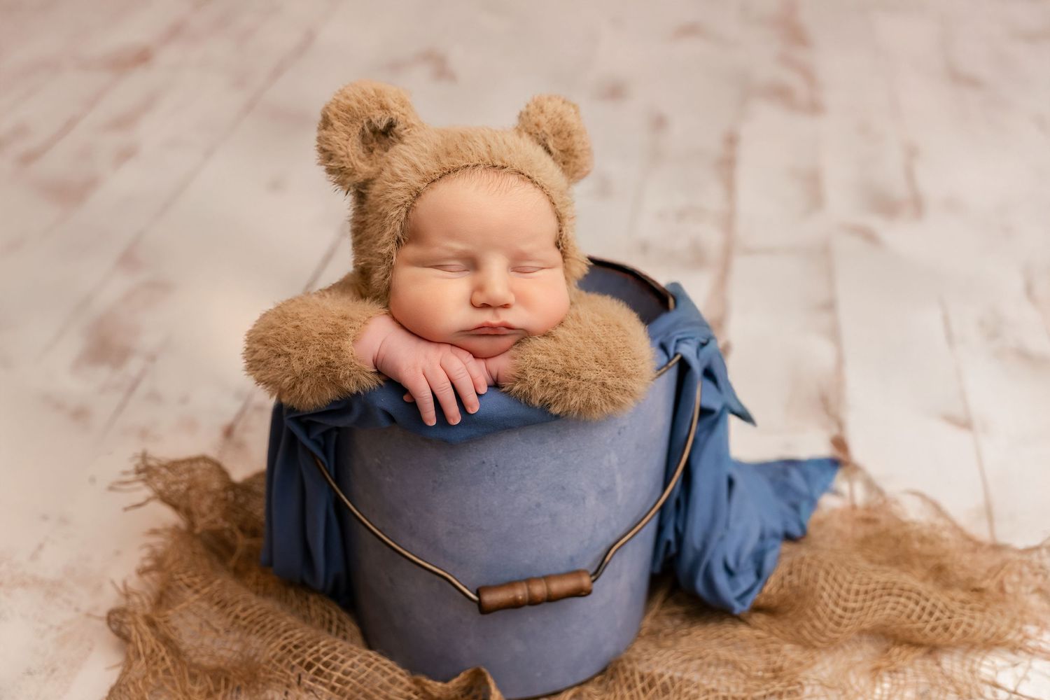 Sleeping newborn baby wearing a brown teddy bear bonnet hat sitting inside a blue bucket on burlap fabric.