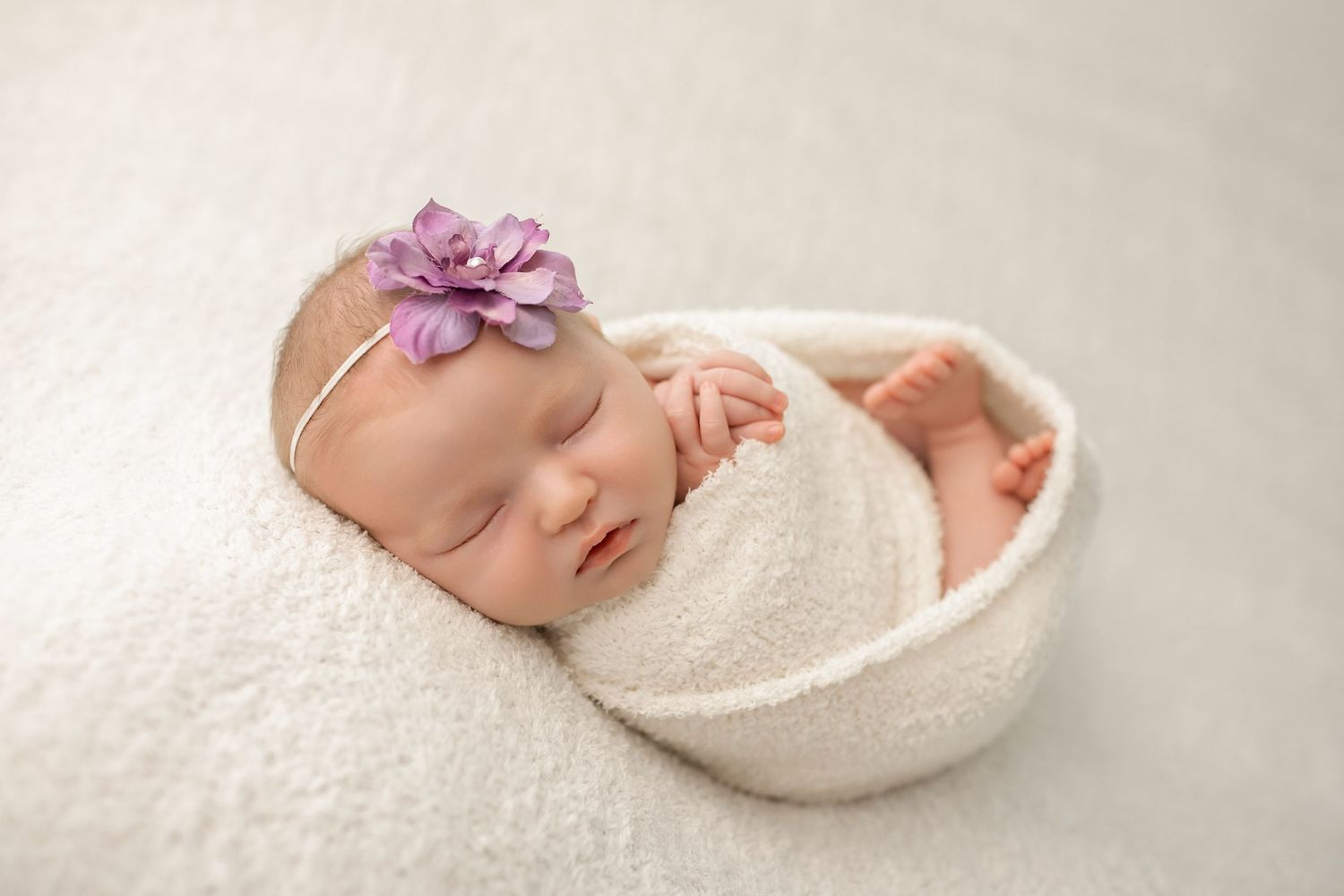 Sleeping newborn baby girl wrapped in white swaddle with purple flower headband in a bowl prop.