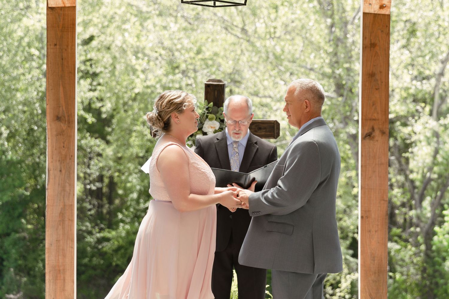 Bride and groom exchange vows outdoors under wooden arch with officiant in lush green garden wedding ceremony.