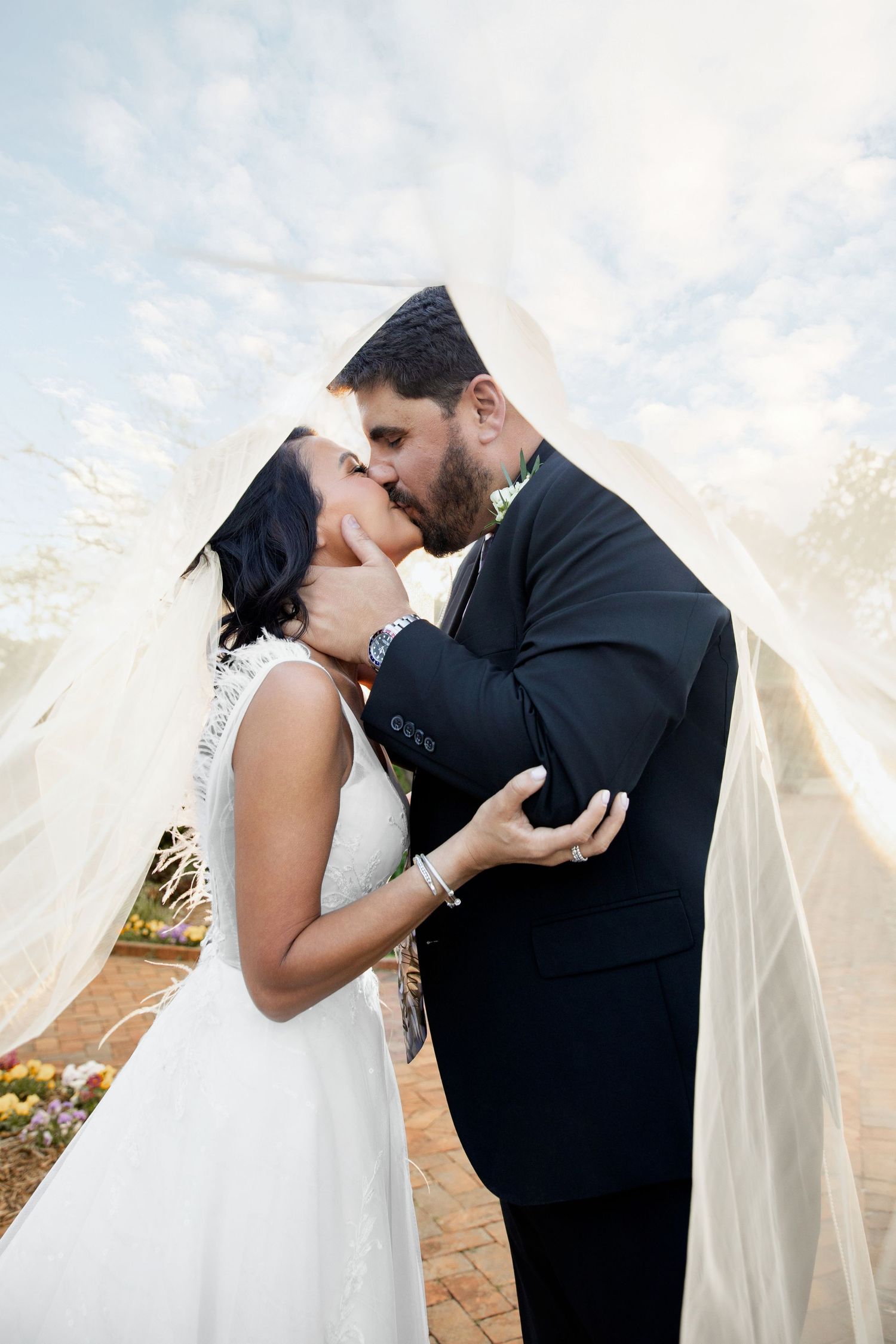 Bride and groom sharing a romantic kiss under a flowing veil on their wedding day outdoors.