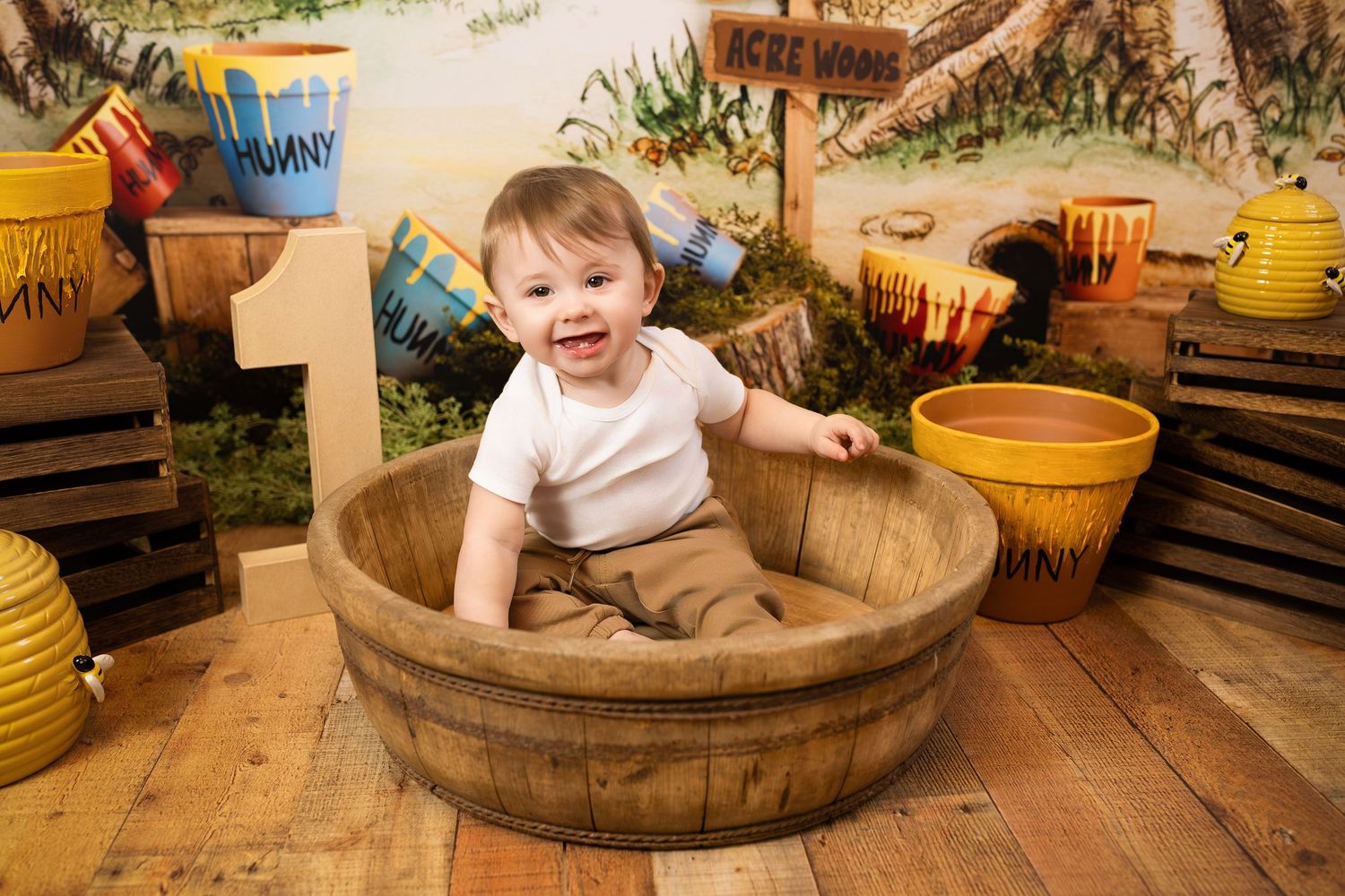 A small child sits happily in a wooden barrel surrounded by yellow ceramic pots on a rustic wooden floor.