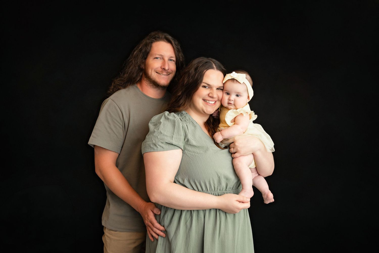 Family portrait of parents and baby in green outfits against black background.
