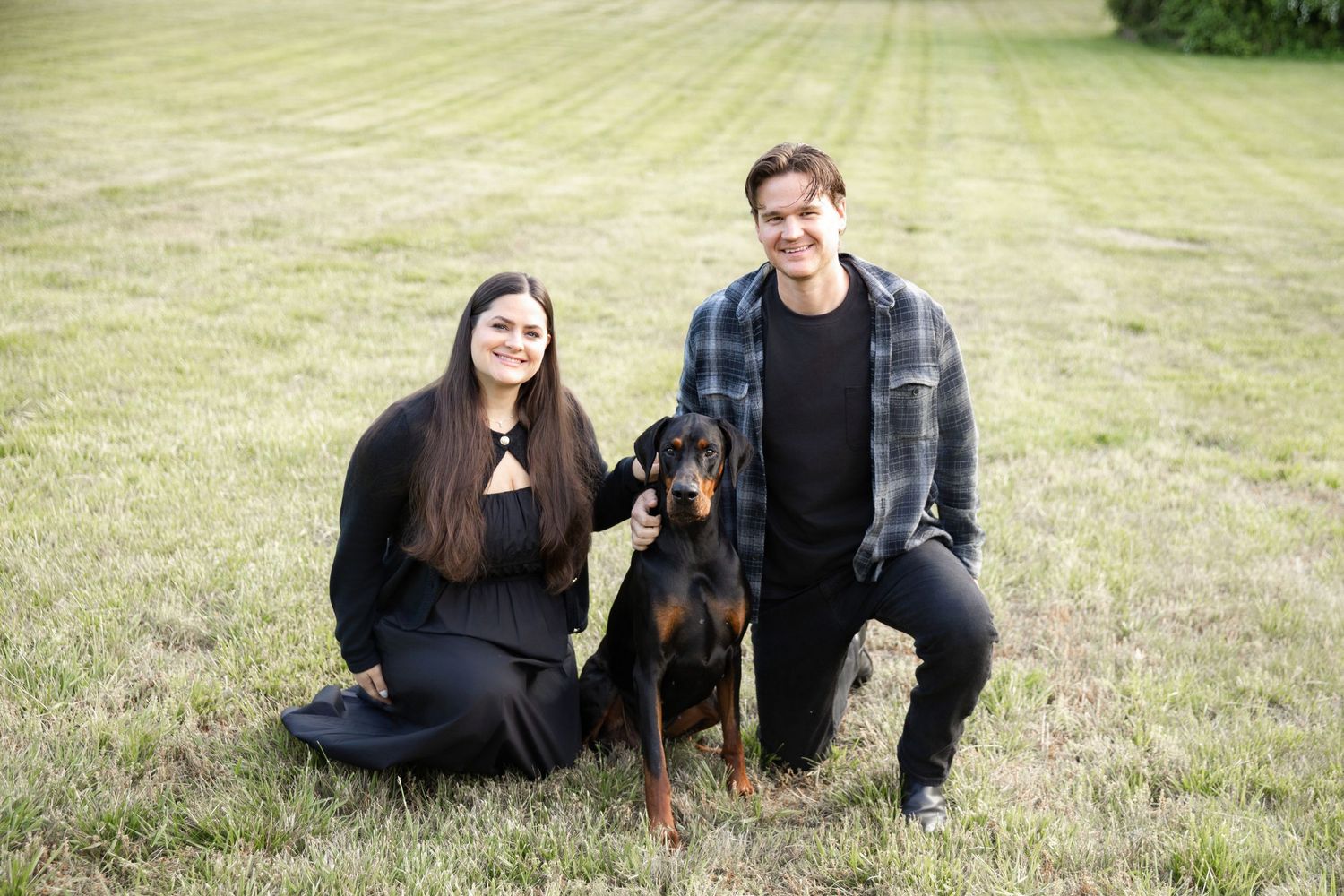 A couple kneeling on green grass with a Doberman Pinscher dog between them outdoors.
