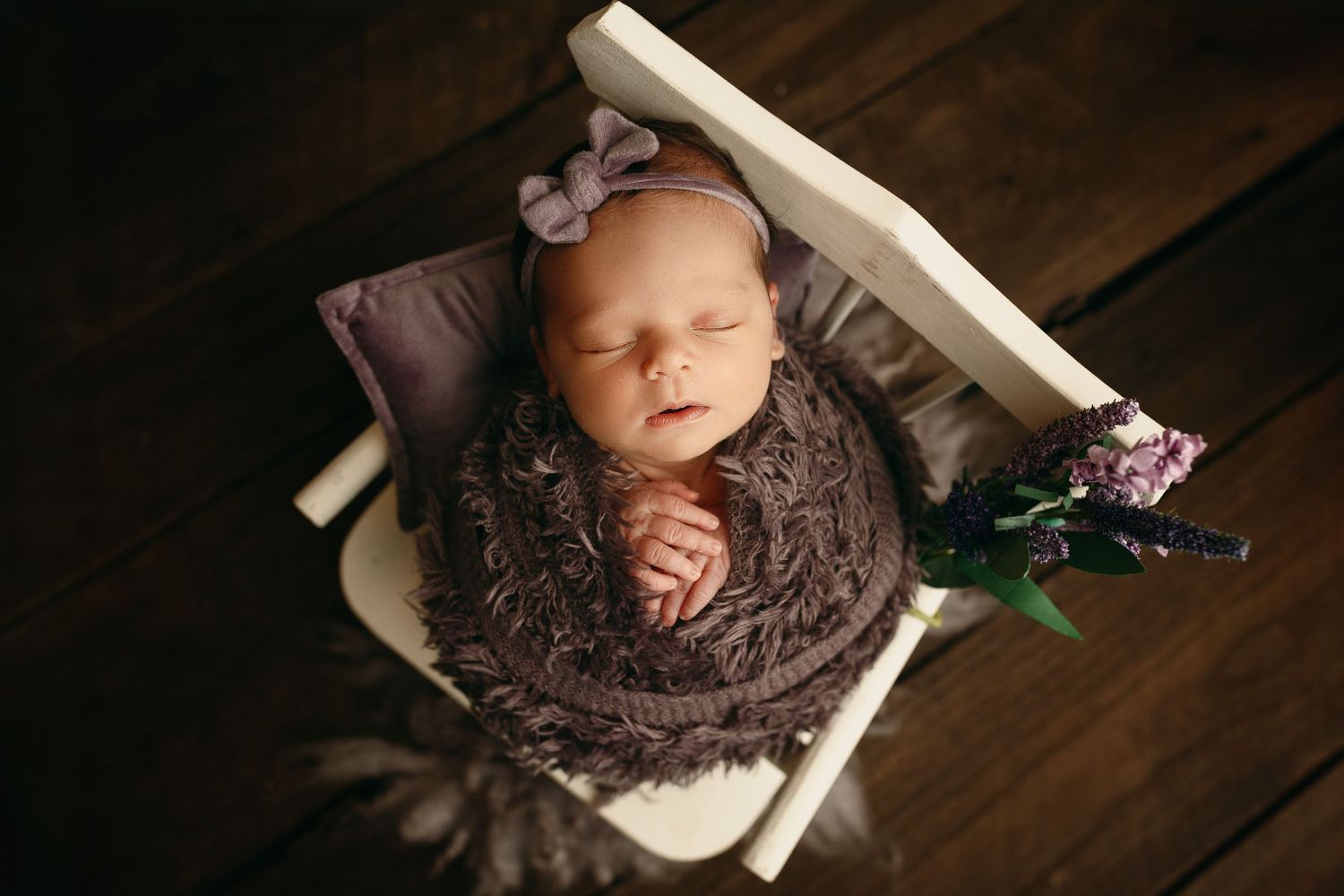 Sleeping newborn baby girl in a purple headband nestled in a knit brown wrap inside a small white wooden chair with lavender flowers.