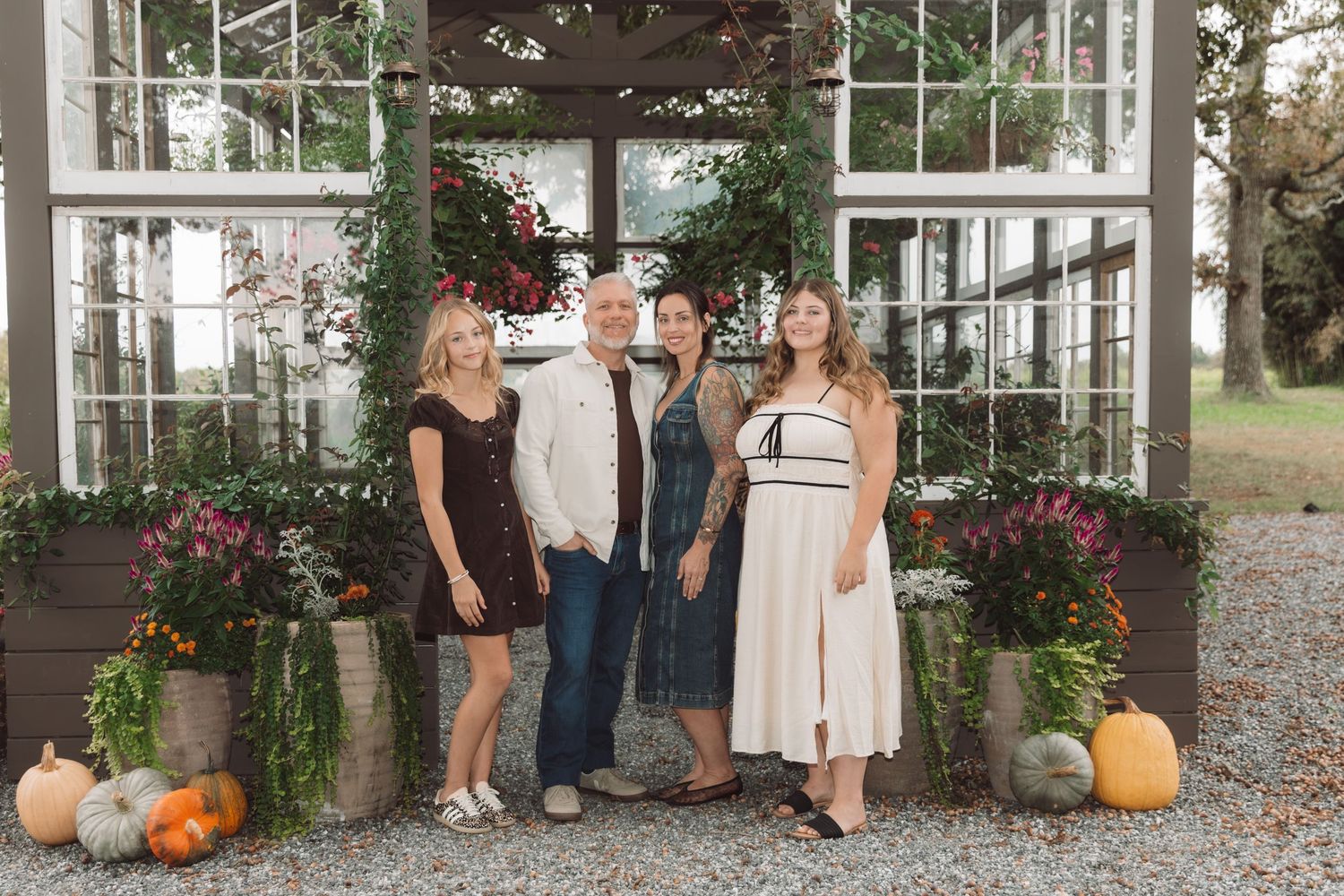 Four people pose together in front of a rustic greenhouse decorated with autumn pumpkins, gourds, and colorful fall flowers.