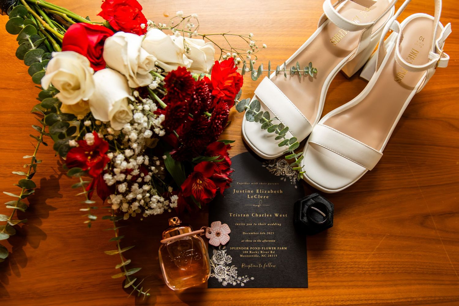 White wedding heels, red and white floral bouquet, black invitation card, and gold perfume bottle arranged on wooden surface.