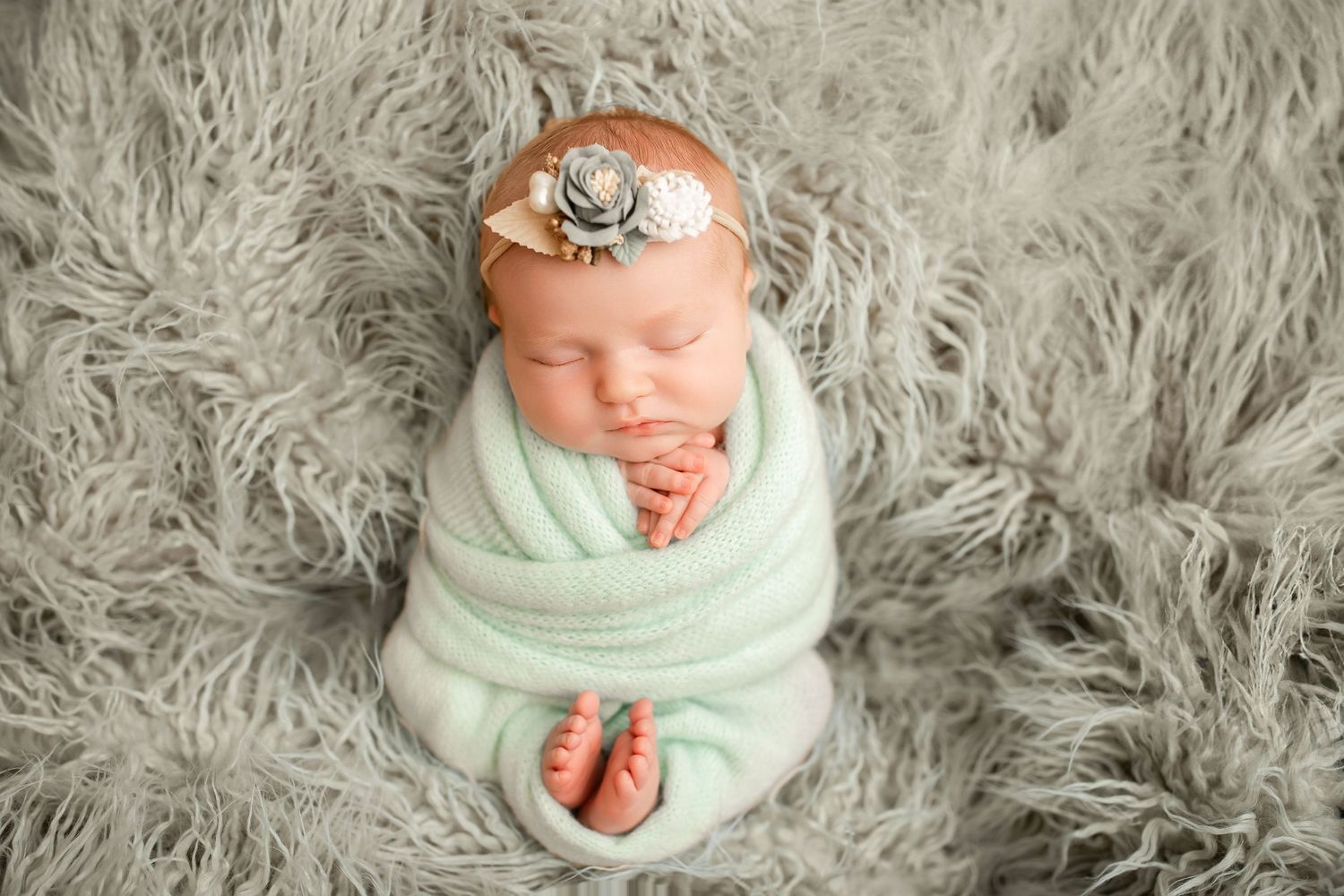 Sleeping newborn baby swaddled in mint green wrap with floral headband on gray fluffy blanket.