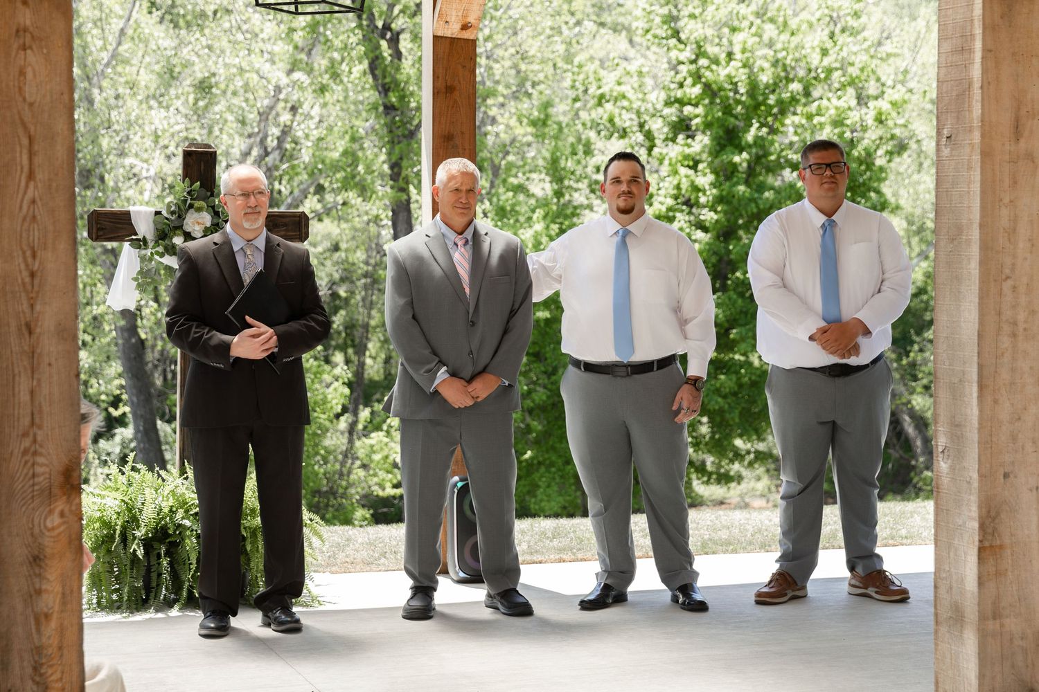Four men in formal attire standing in an outdoor pavilion surrounded by lush green trees at a wedding ceremony.