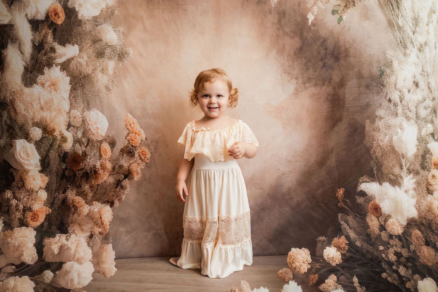 A smiling toddler girl in a cream dress stands before a floral arch backdrop with soft peach and white blooms.