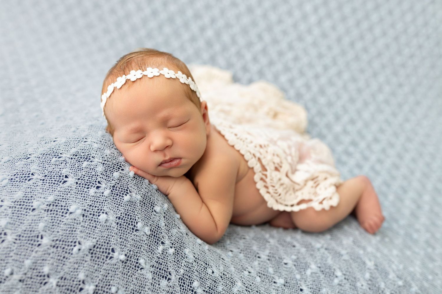 Newborn baby girl sleeping peacefully on blue blanket wearing lace wrap and floral headband in professional photo.