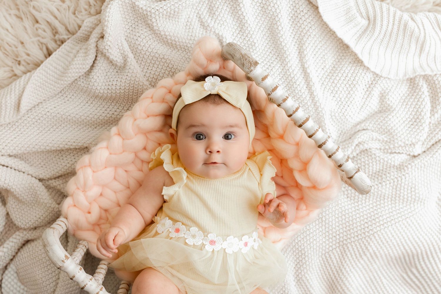 Baby girl in a pale yellow dress and matching bow headband lying on a cozy knit blanket with a peach chunky yarn wrap and wooden beads.