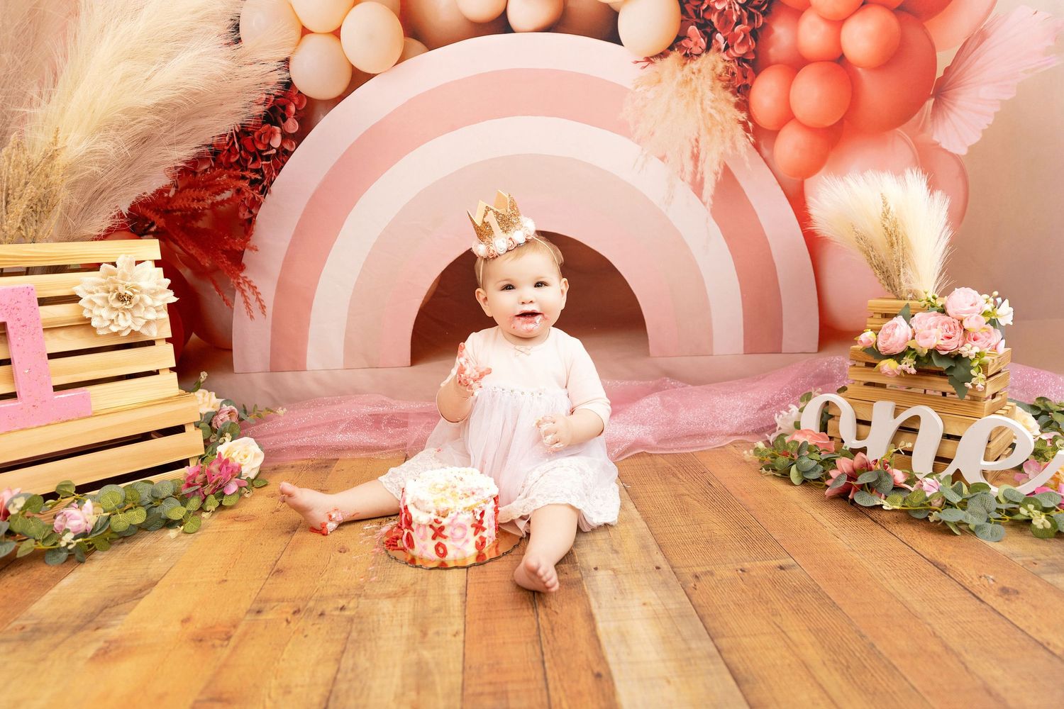 Pink and peach balloon arch decorates a first birthday photoshoot setup with floral accents and vintage props.