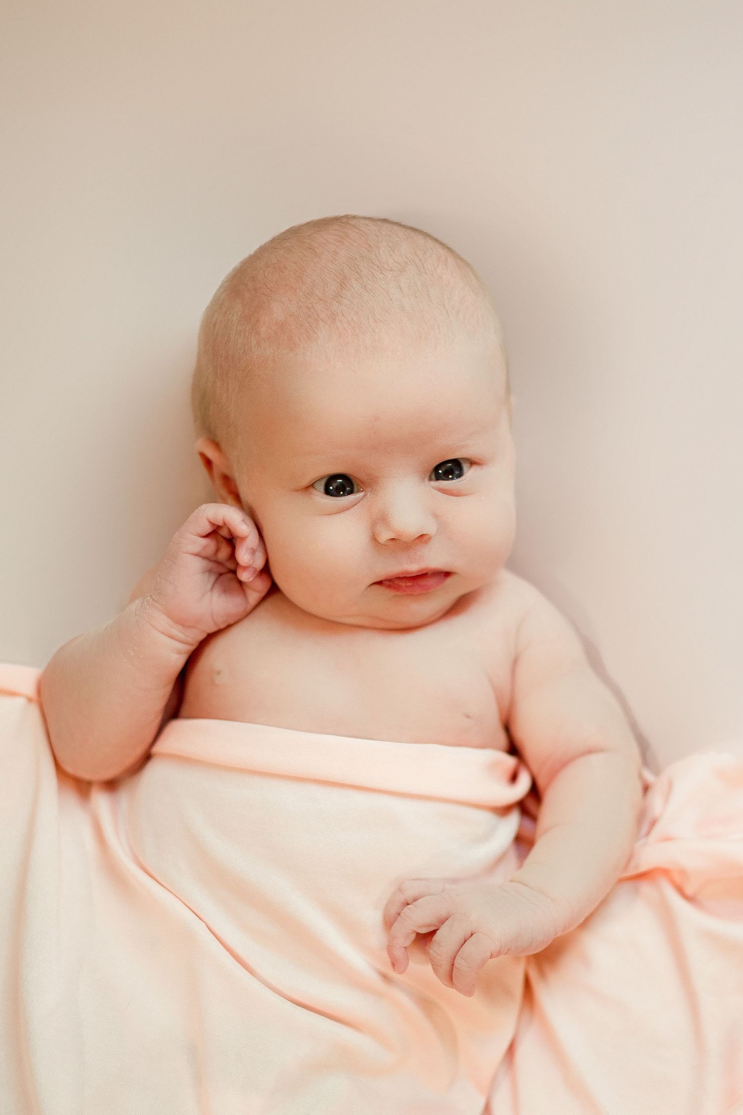 Sweet baby wrapped in soft peach blanket touching ear while looking at camera against neutral background.
