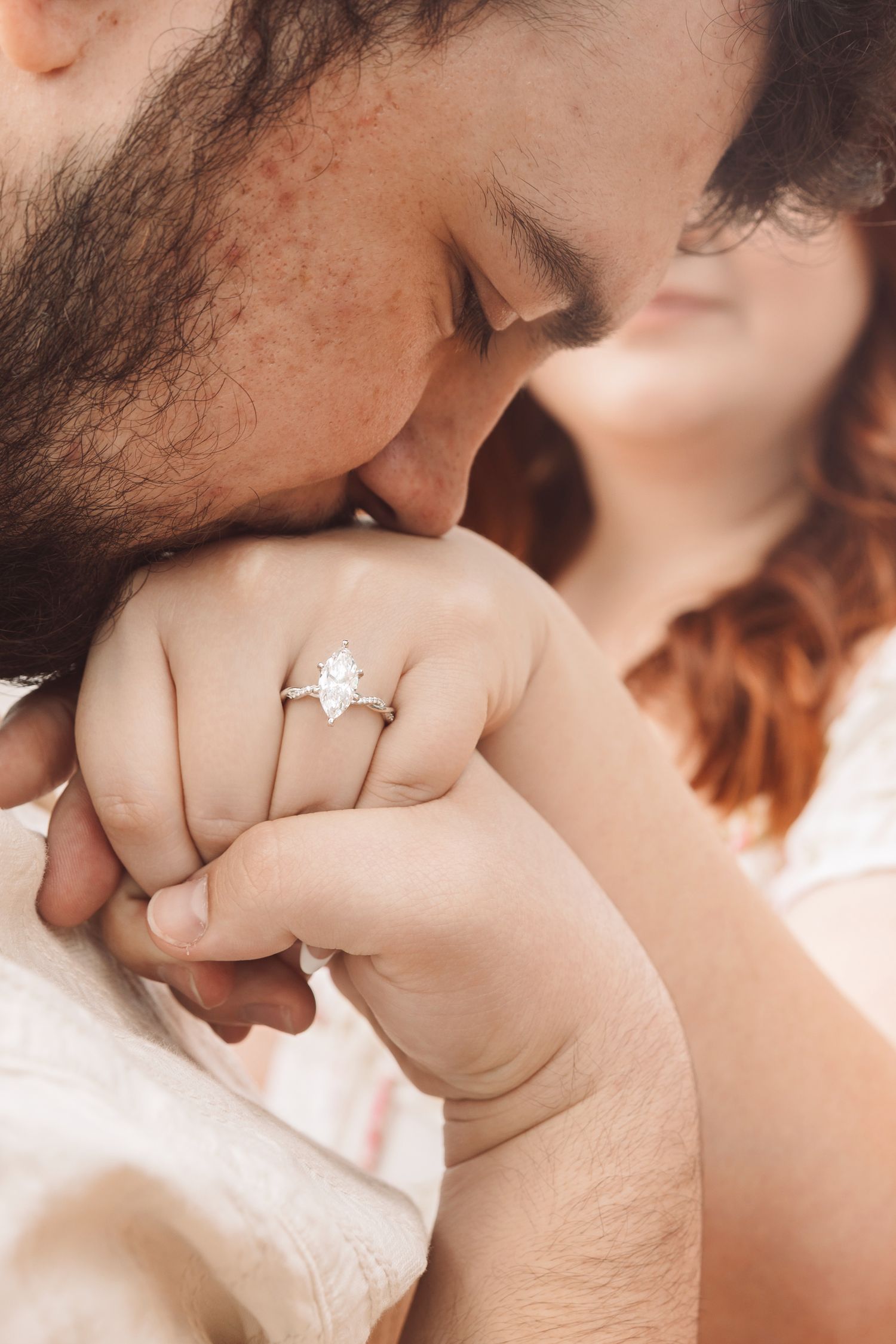 A man kisses a woman's hand adorned with a sparkling pear-shaped diamond engagement ring in a romantic close-up.