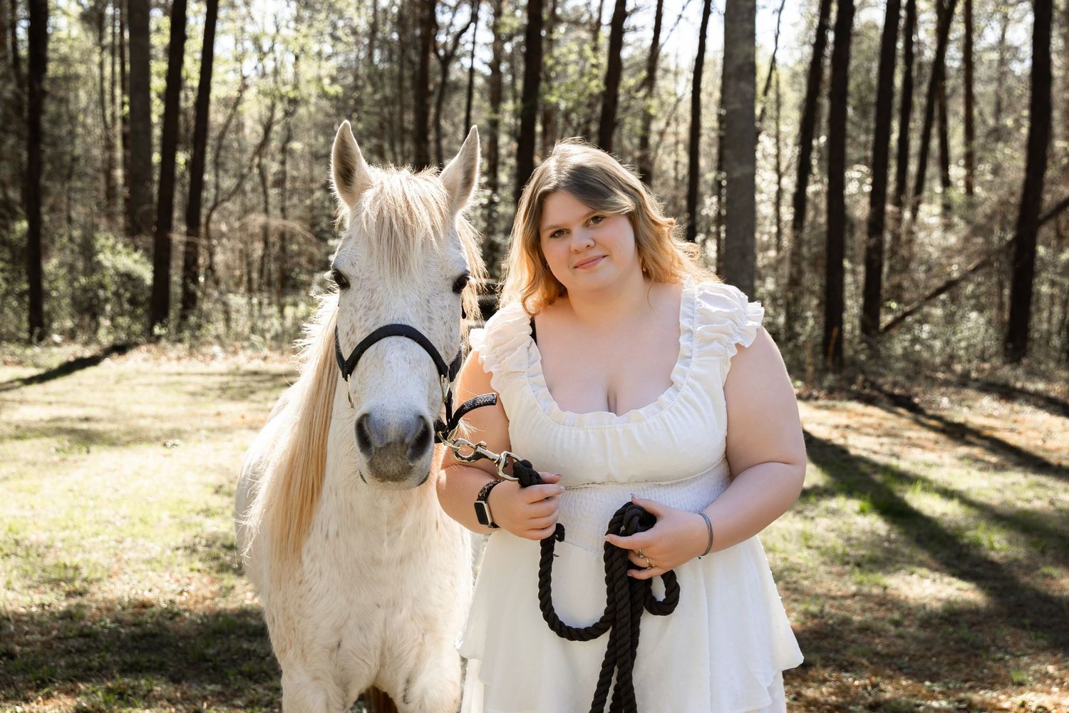 Woman in white dress stands beside a white horse on a lead rope in a sunlit forest clearing.