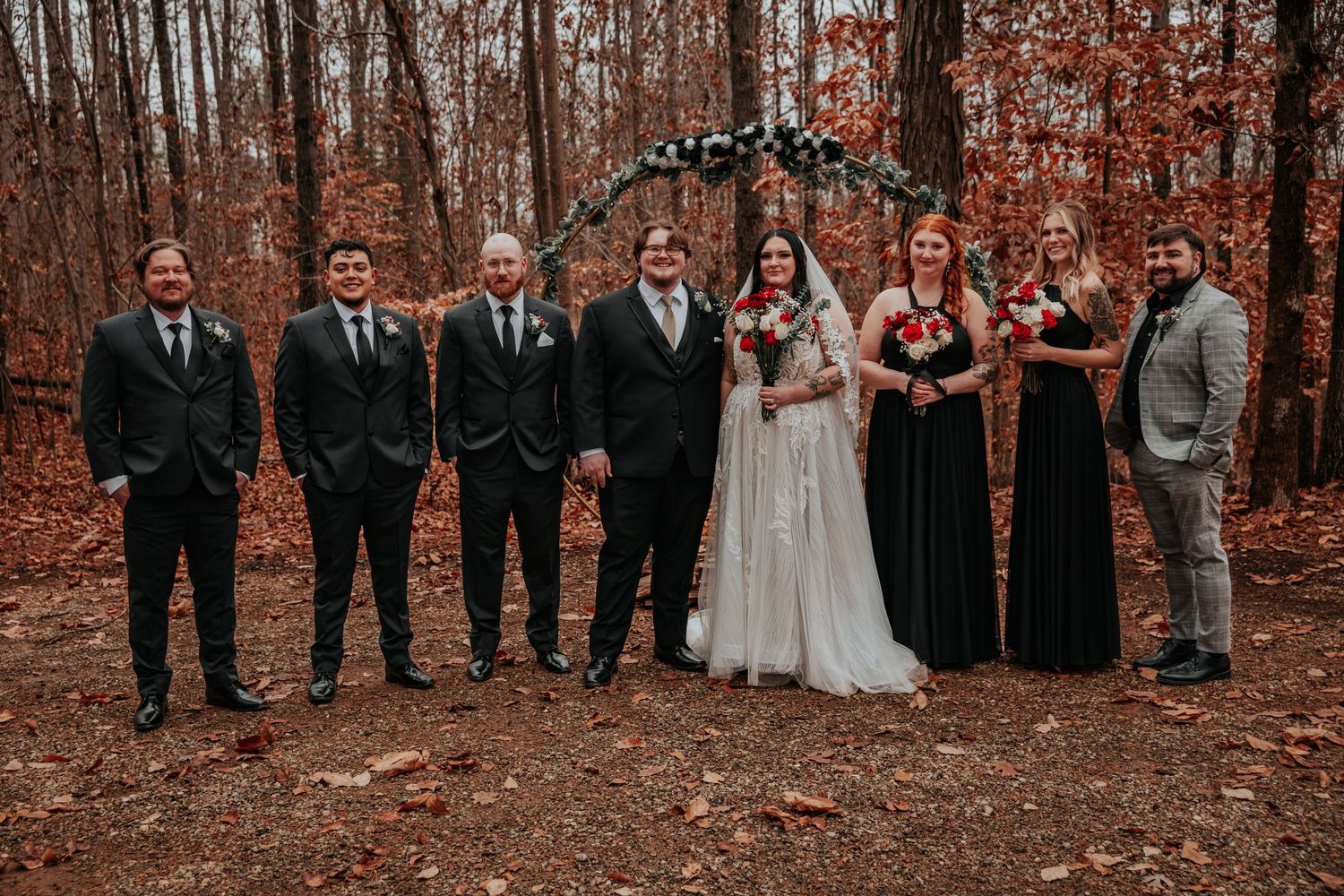 Wedding party poses in autumn forest with couple and attendants in black formal wear.