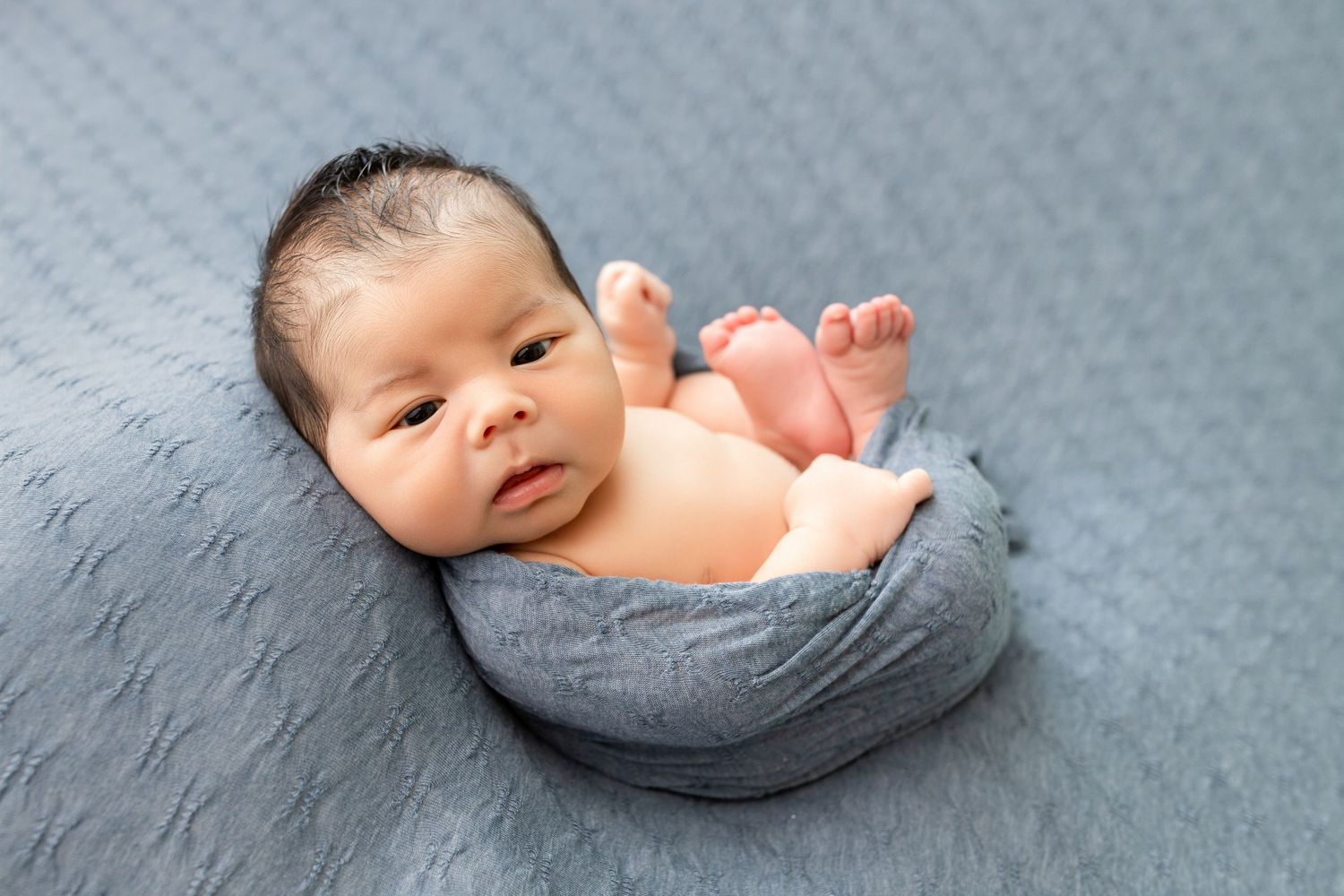 Newborn baby wrapped in gray cloth lying on blue textured blanket with feet raised.