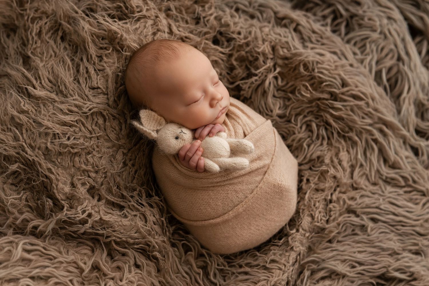 Newborn baby swaddled in beige wrap holding a small bunny toy, sleeping peacefully on a brown fluffy blanket.