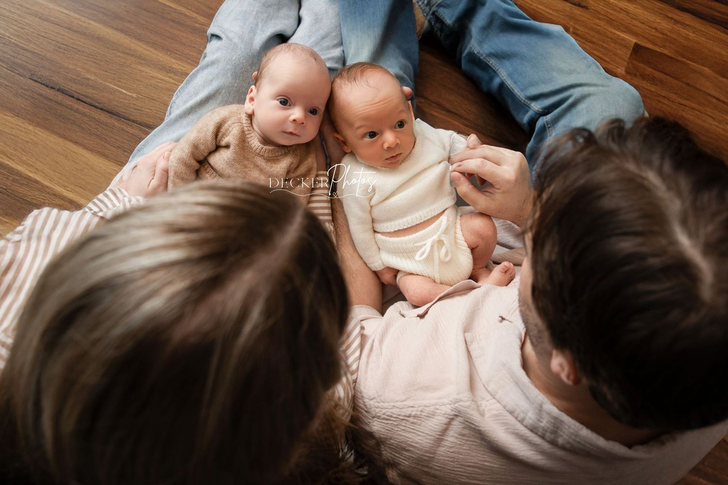 Two parents sitting on hardwood floor with twin babies between them, viewed from above in a cozy home setting.