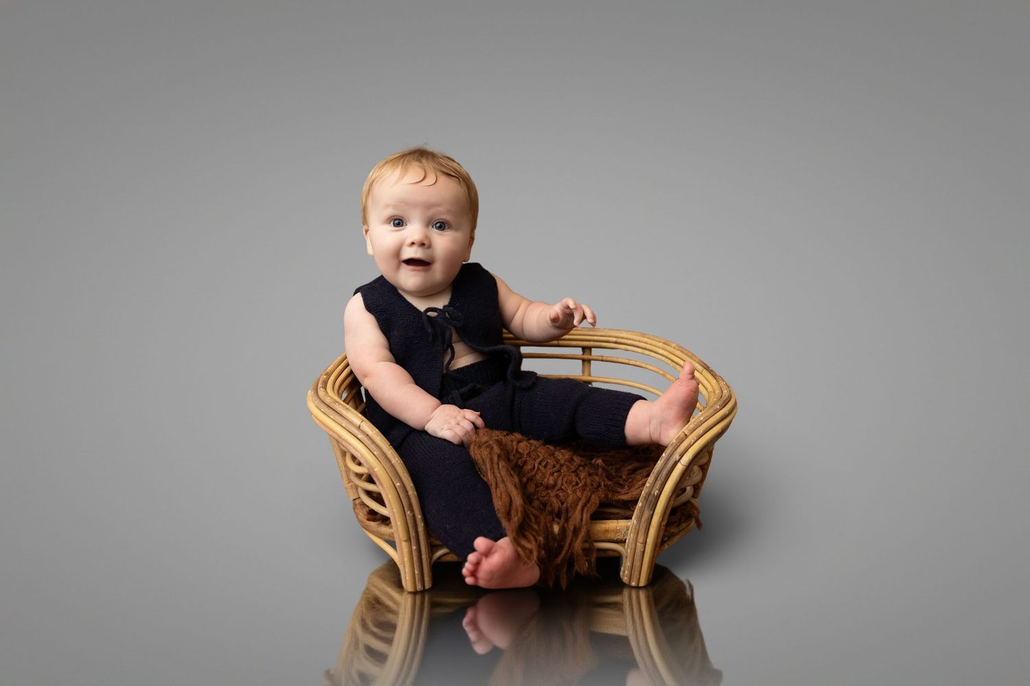 A smiling baby in a navy romper sits in a small wicker chair on a gray studio background.