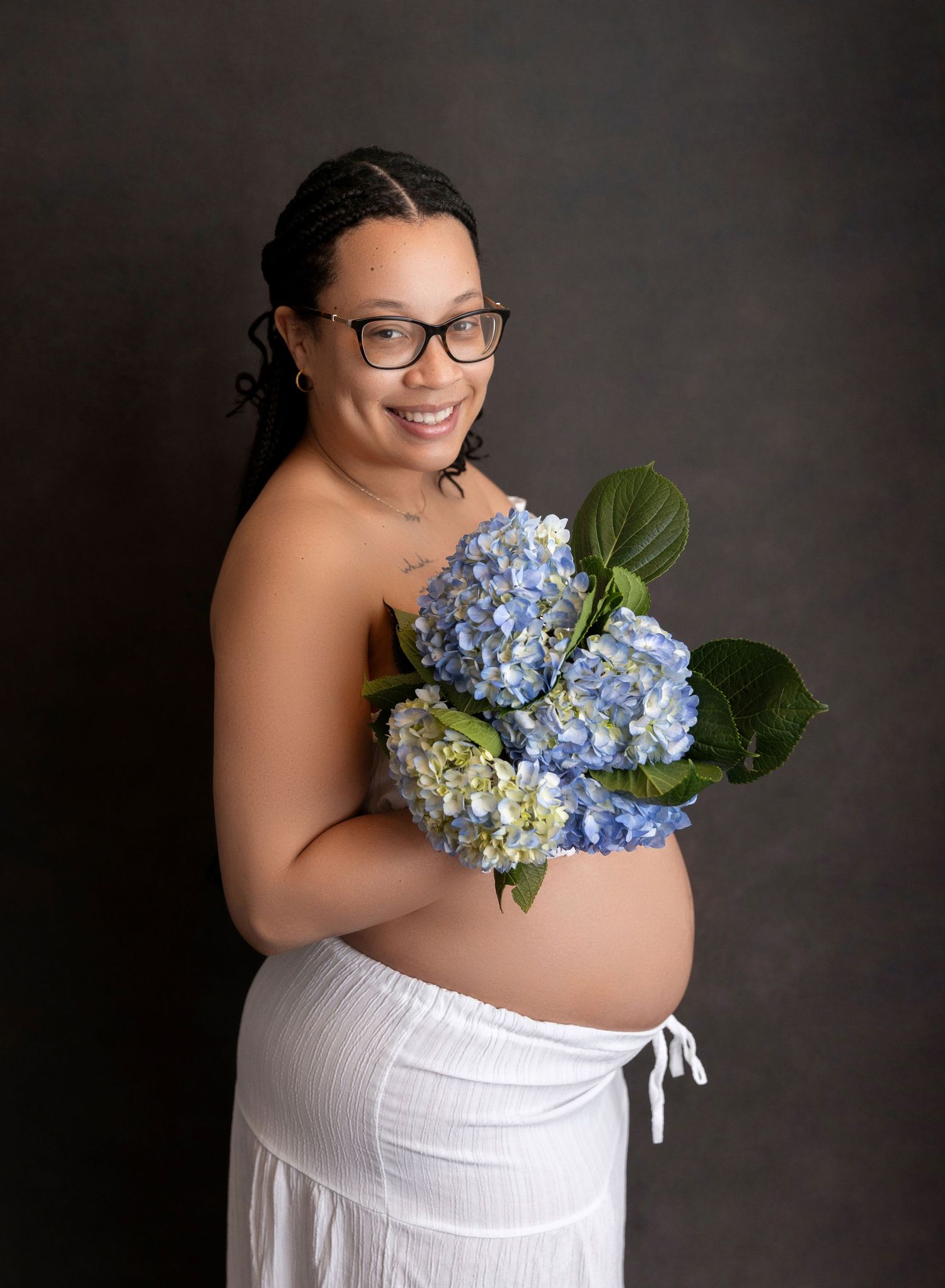 Smiling pregnant woman in white skirt holding blue hydrangeas against dark background.