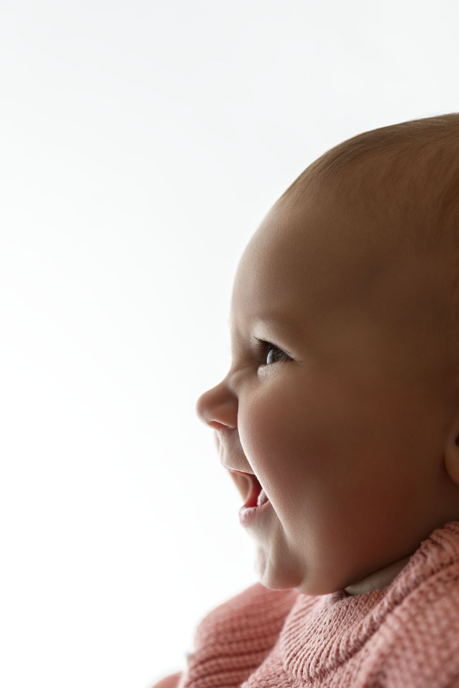 Side profile of a smiling baby in a pink knit sweater against a bright white background, mouth open in joyful laughter.