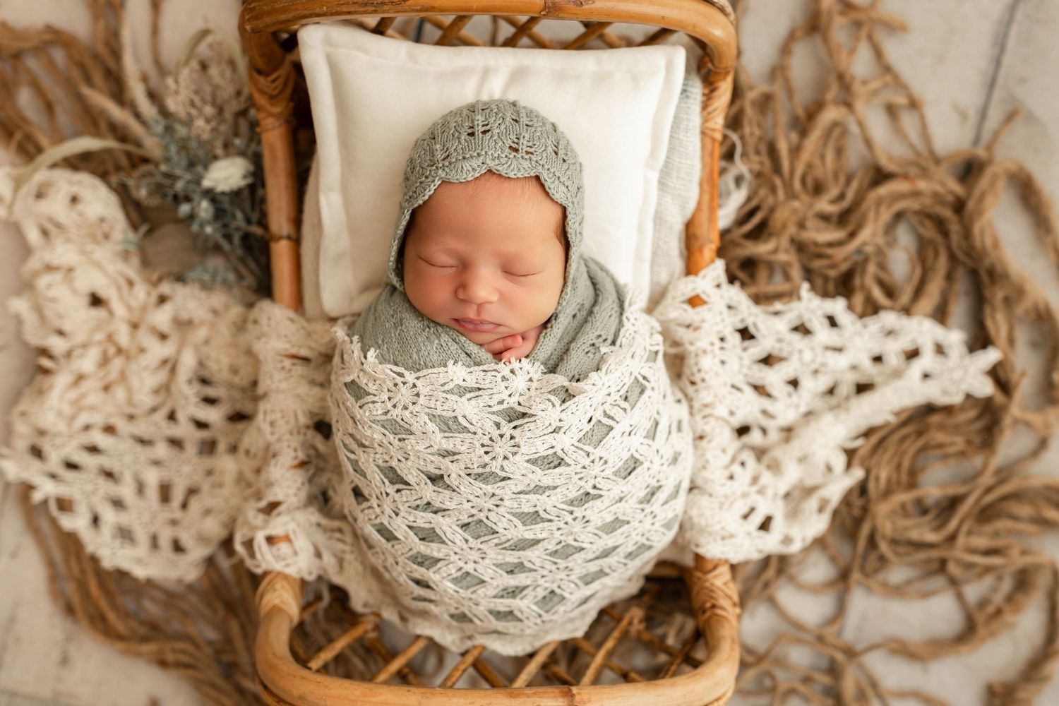 Newborn baby swaddled in sage green knit wrap and bonnet, sleeping peacefully in a wicker basket.